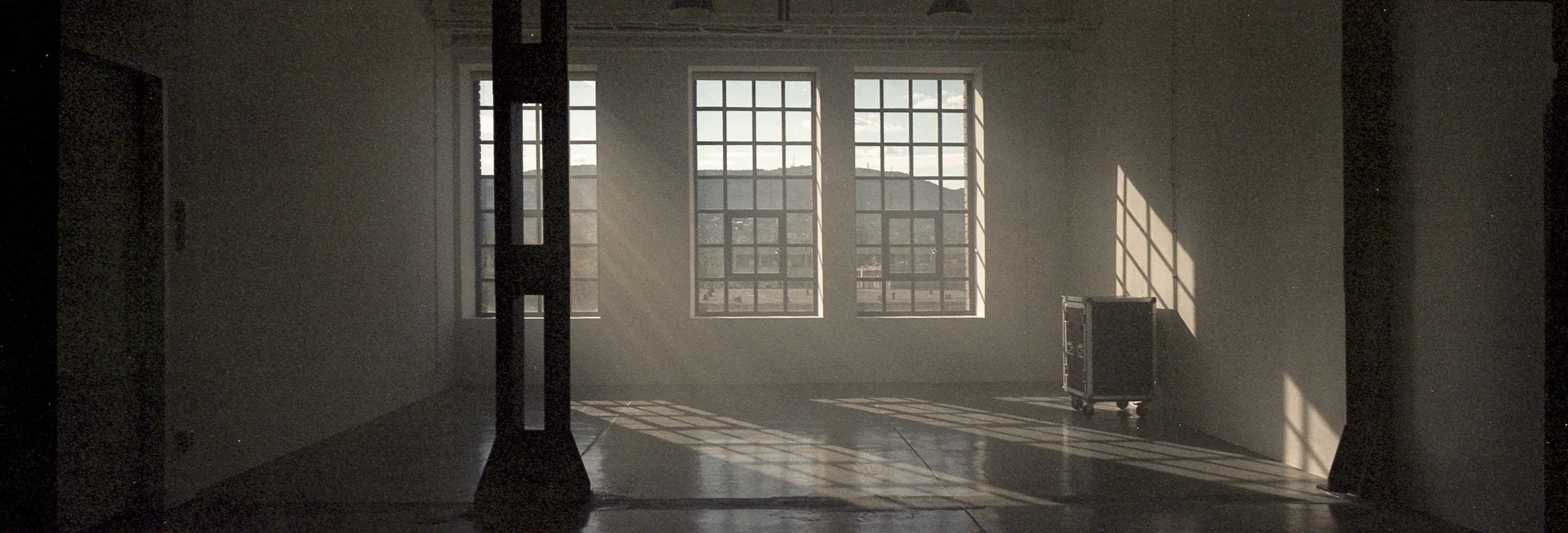 An empty room with large windows letting in sunlight, casting shadows on the floor, and a small cart near the wall.