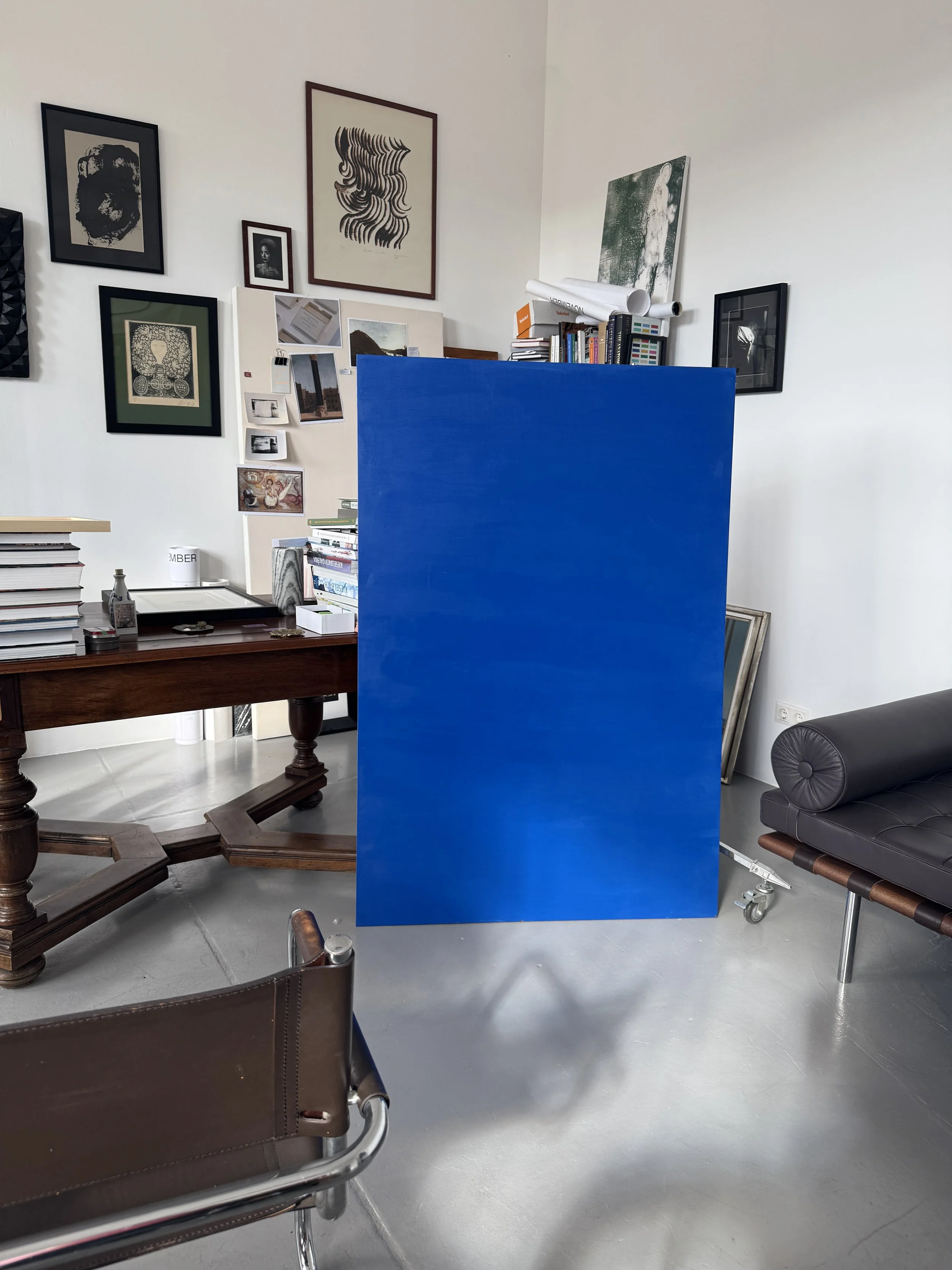 Interior of an art studio or gallery with various framed artworks on white walls, a wooden table with books and art supplies, a black leather couch, and a bright blue panel in the center.