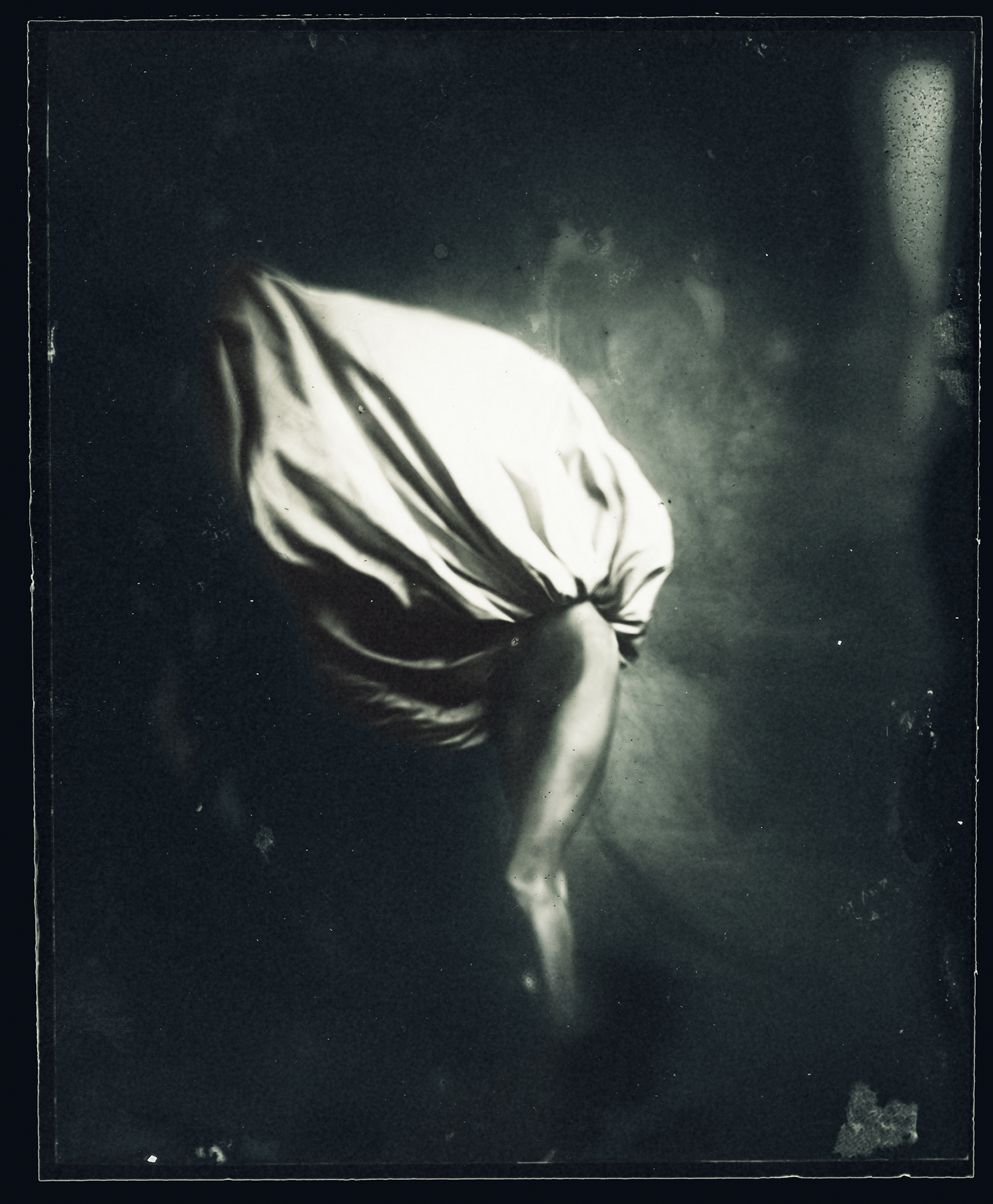 A person sitting with their head bowed and hands covering their face, draped in a white cloth, in a dimly lit setting.