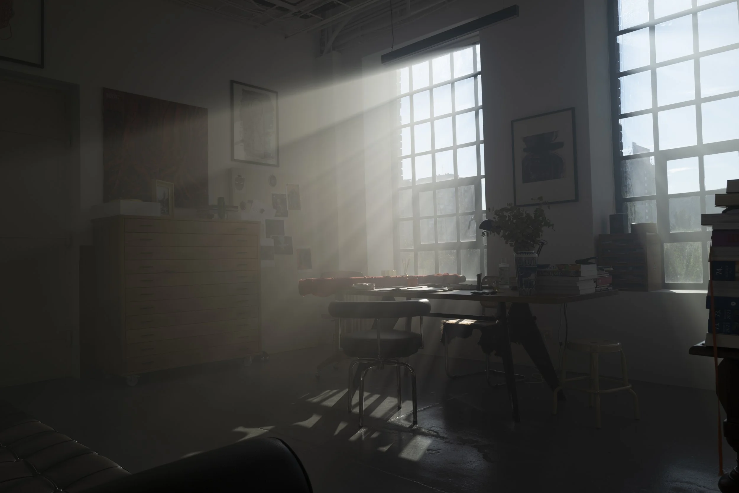 Sunlight streaming through large windows into an artist's studio or office space with furniture, books, and artwork on the walls, creating a bright and airy atmosphere.
