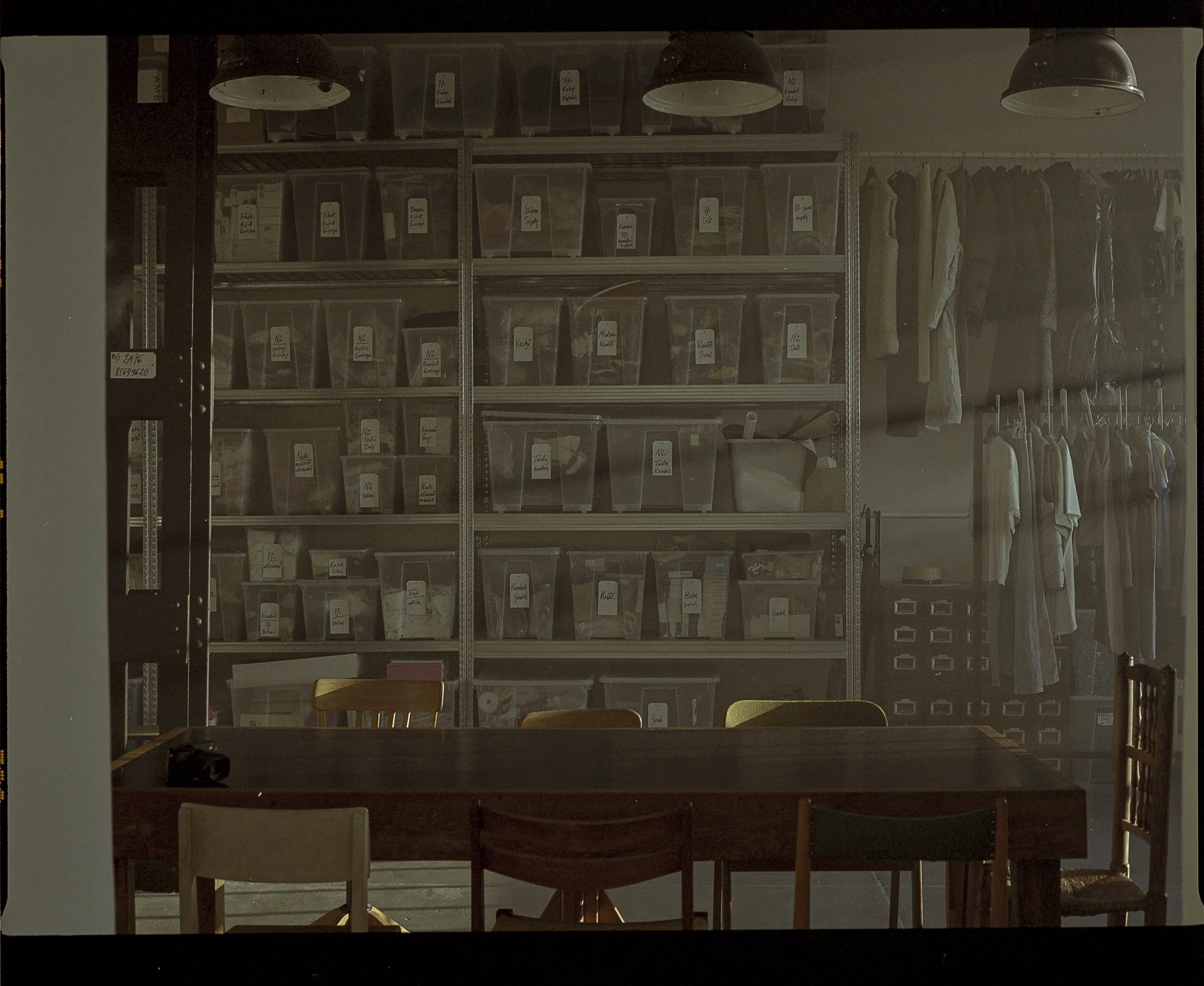A room with shelves filled with labeled plastic storage bins, a table with chairs in front, and hanging clothes on the right side.