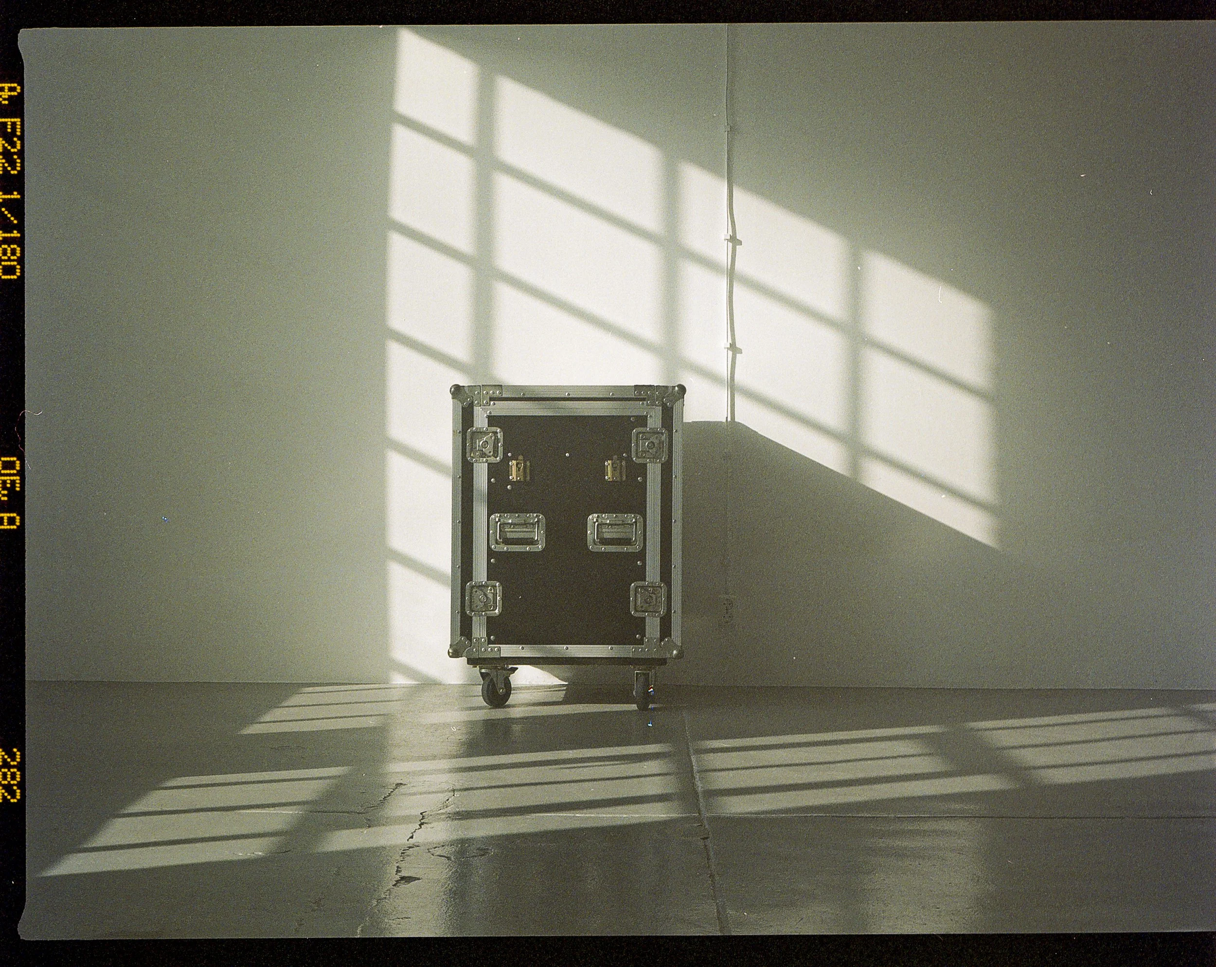 A black storage or transportation case on wheels with metal framing, placed against a plain white wall, with shadows cast by nearby window panes creating a grid pattern on the wall and floor.