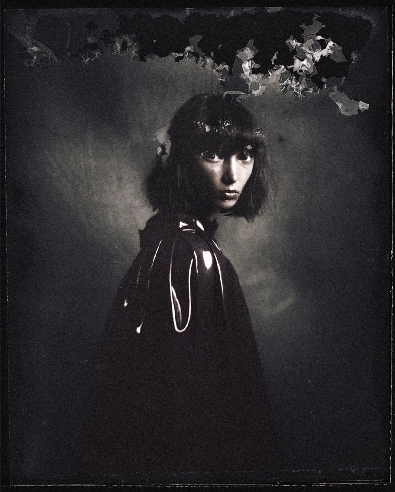 A black and white portrait of a woman with shoulder-length dark hair and bangs, wearing a shiny dark coat, looking over her shoulder with a serious expression, with leaves and abstract shapes above her head.