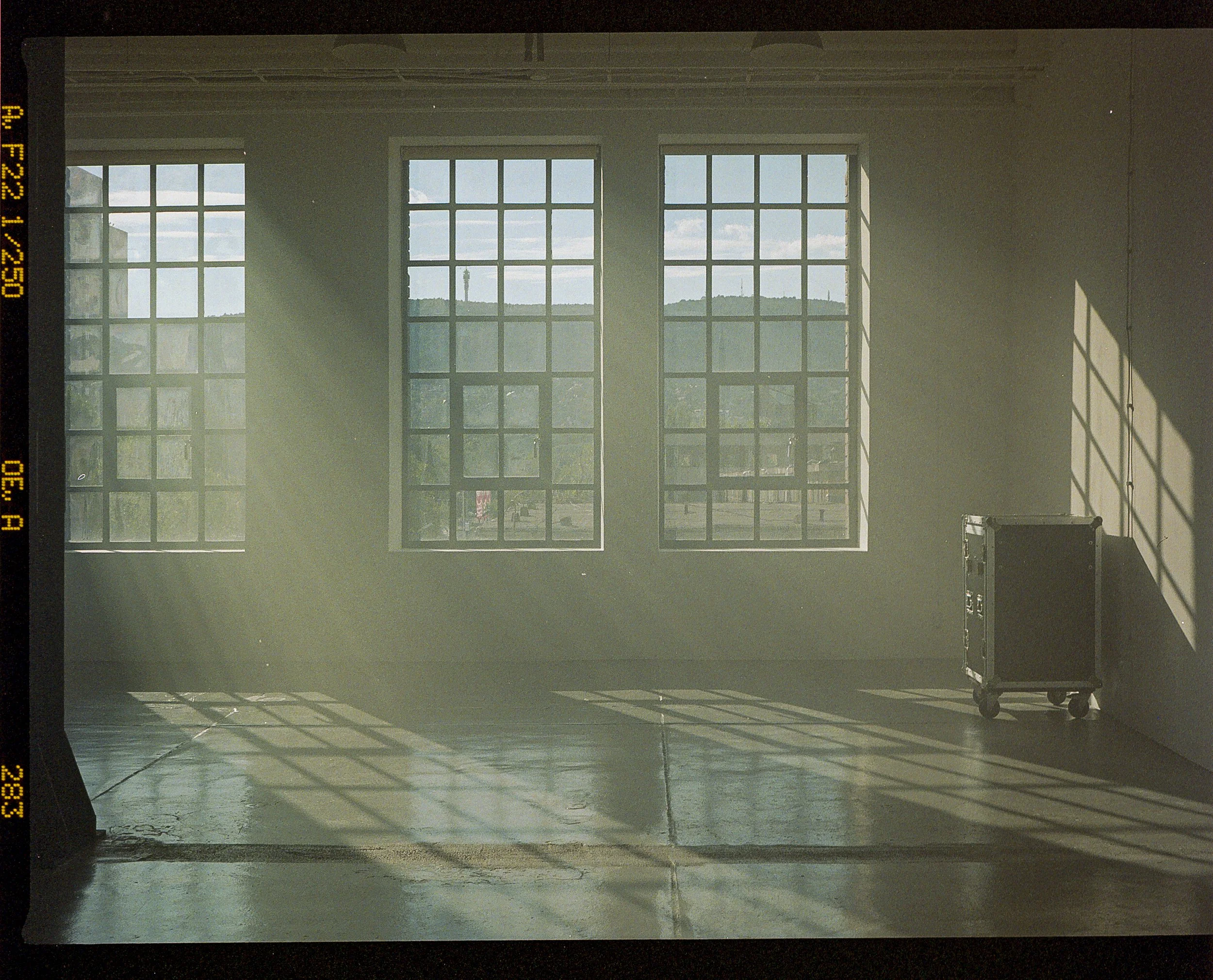 Sunlight streams through three large grid-pattern windows into a sparse, empty room with a smooth floor. A small rolling black case is positioned near the wall on the right.