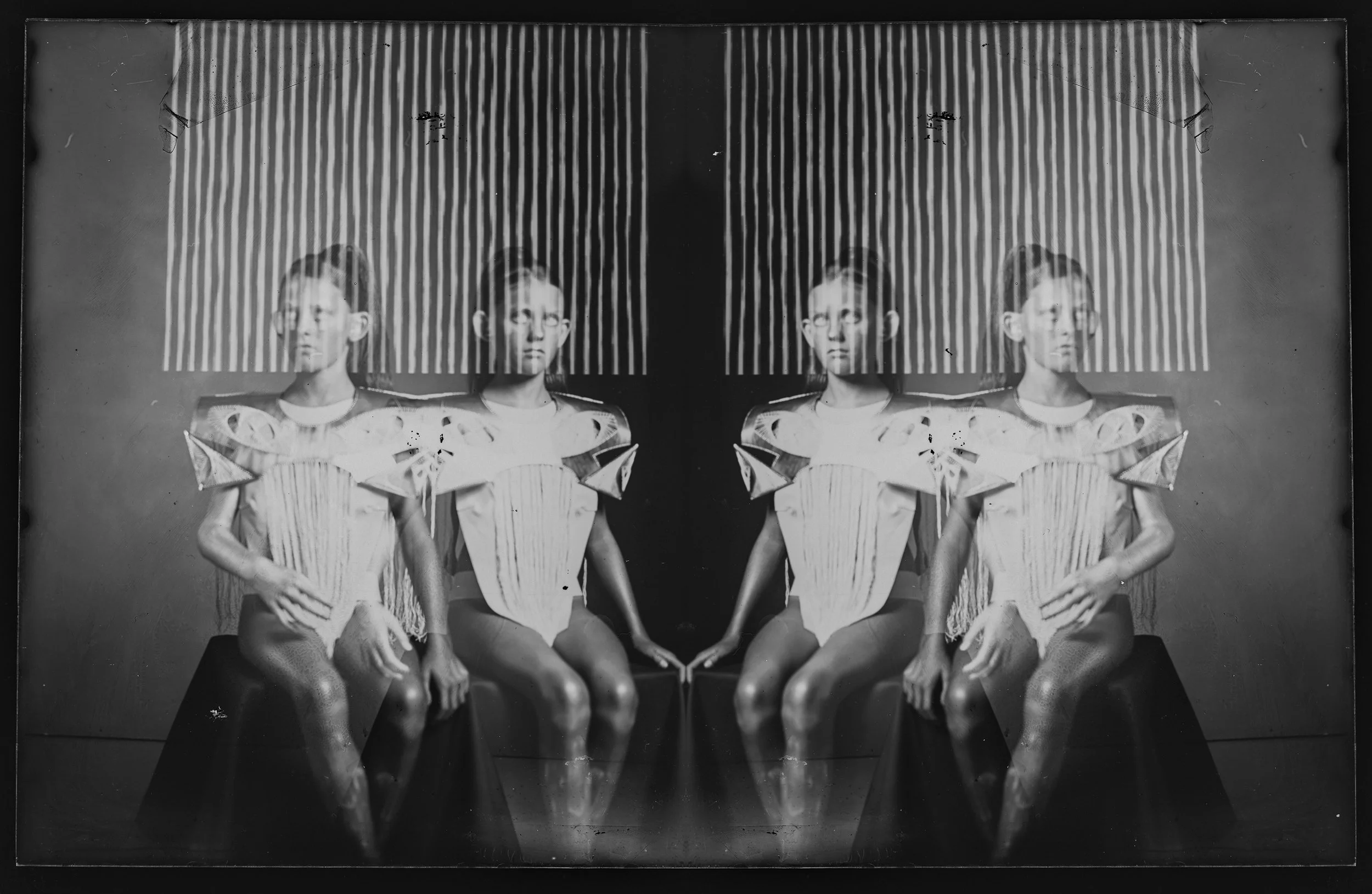 Black and white photograph of a woman duplicated to create a mirrored image, sitting on a bench with a black background and vertical light stripes. She is wearing a futuristic, sculptural dress with exaggerated shoulders.