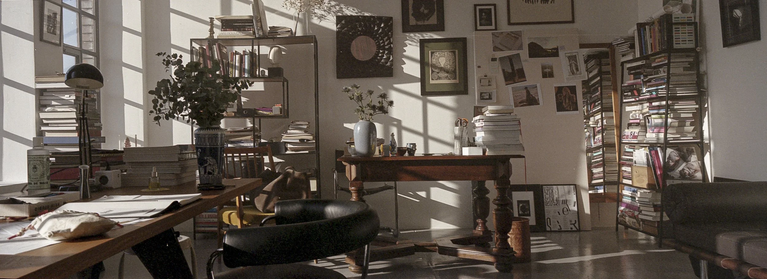 Sunlight streaming through large windows into an overstuffed home office or studio filled with books, papers, and artwork, with a desk, chairs, and various decorative items.