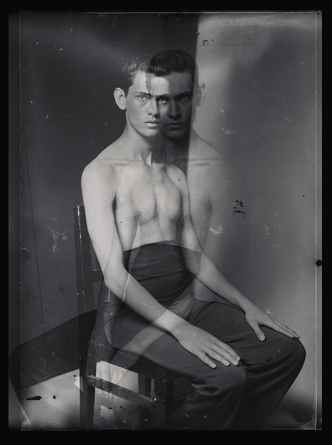 Black and white photograph of a young male model sitting on a chair, with a multiple exposure effect creating a ghostly duplicate. The model is shirtless, wearing dark trousers, with a serious expression, and the background is plain.