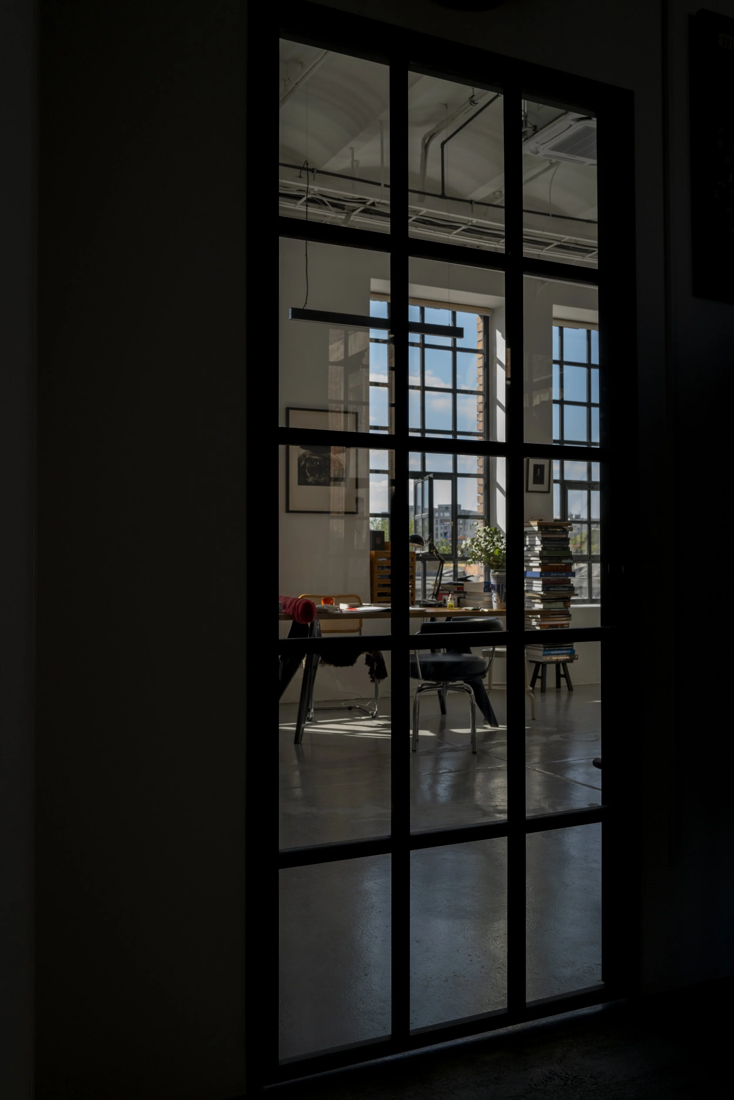 An office space seen through a glass door with grid panes. Inside, large windows let in natural light, and there is a stack of books, a desk with office supplies, and chairs.