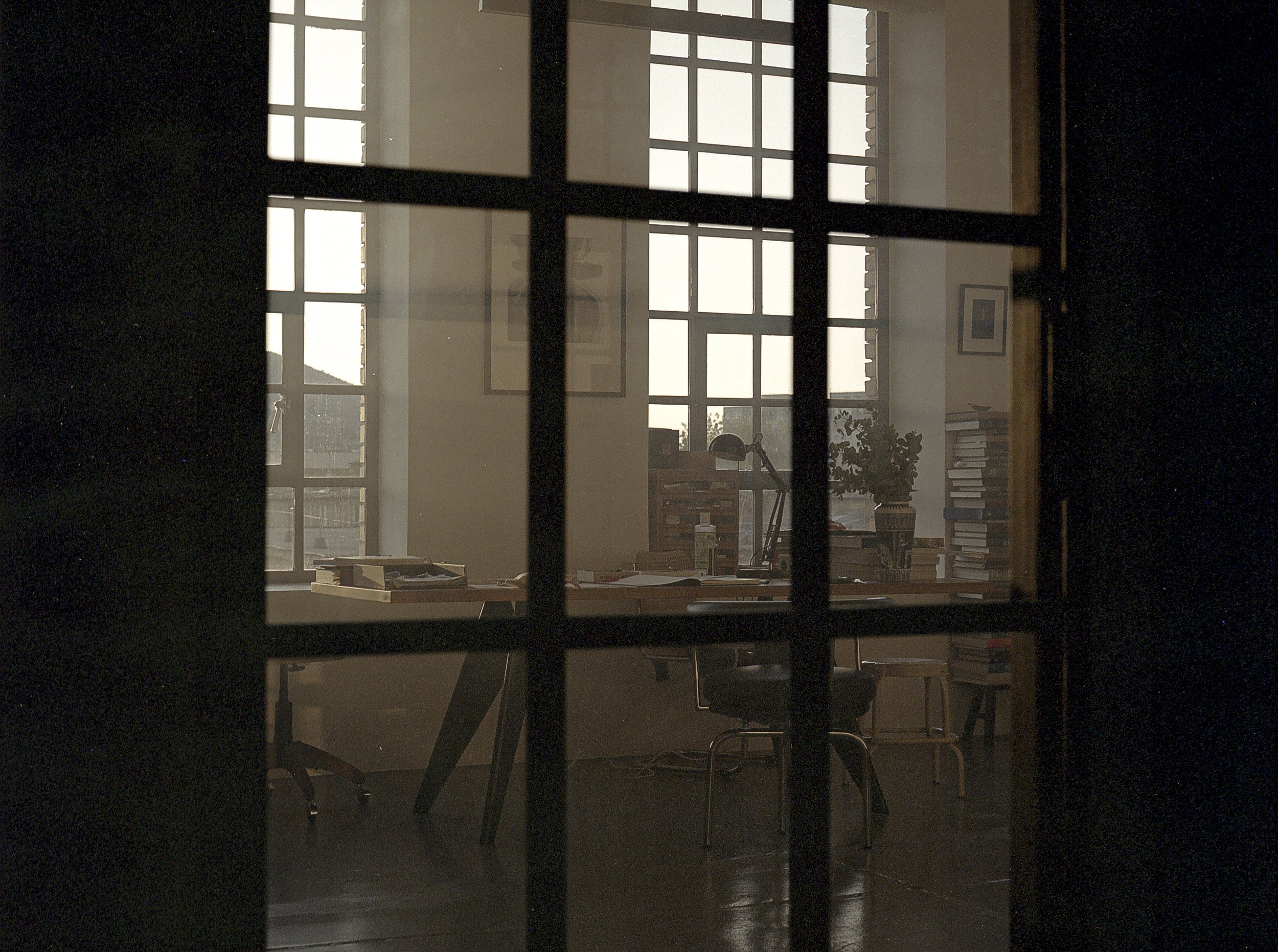 An office seen through a glass-paneled door with a view of a cluttered desk, a chair, a large window, and various potted plants and framed pictures.