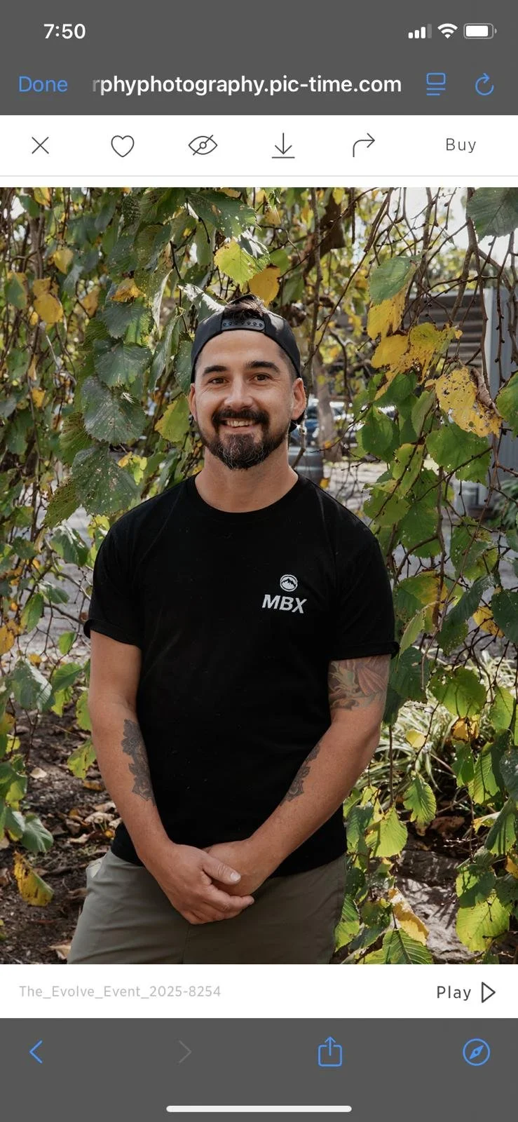 Smiling man with a beard, tattoos on arms, wearing a black T-shirt with 'MBX' logo and a black backward cap, standing outdoors with green and yellow leaves in the background.