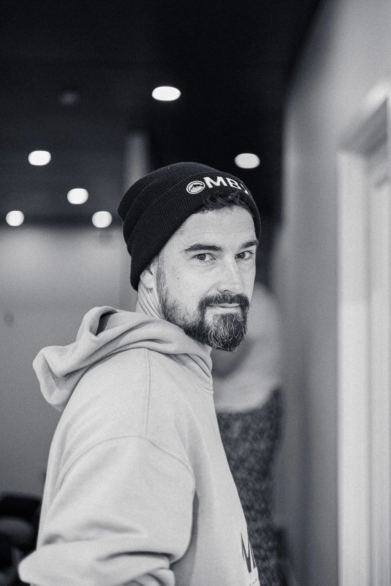 Black and white photo of a man with a beard and mustache wearing a beanie and hoodie, looking over his shoulder at the camera.
