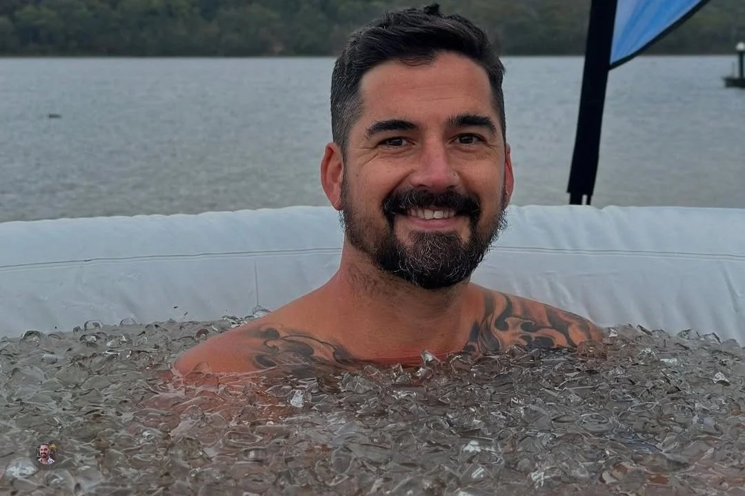A man with a beard and tattoos smiling in a hot tub filled with ice, outdoors near a lake.