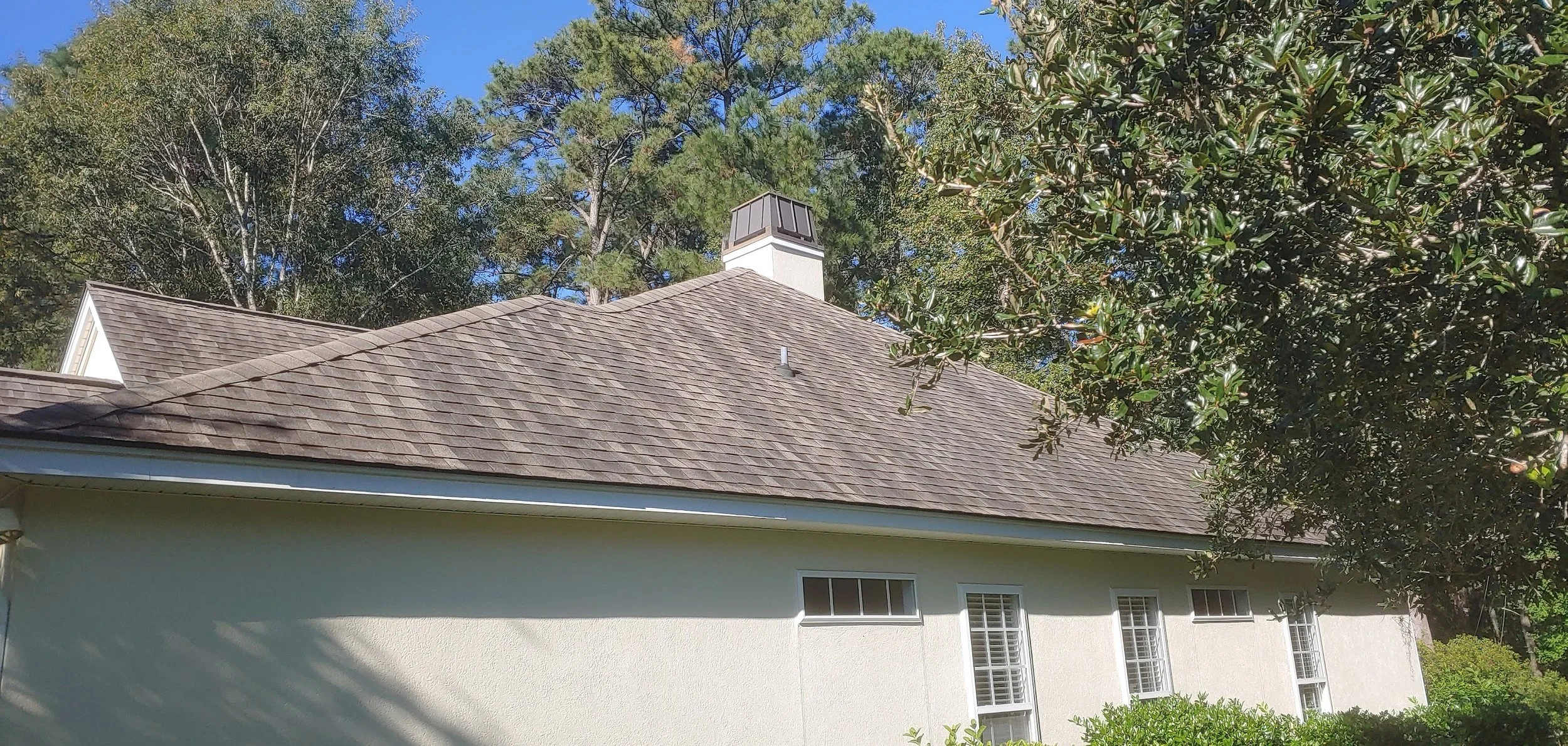 Photo of a house roof with shingles, surrounded by trees and greenery, under a clear blue sky.
