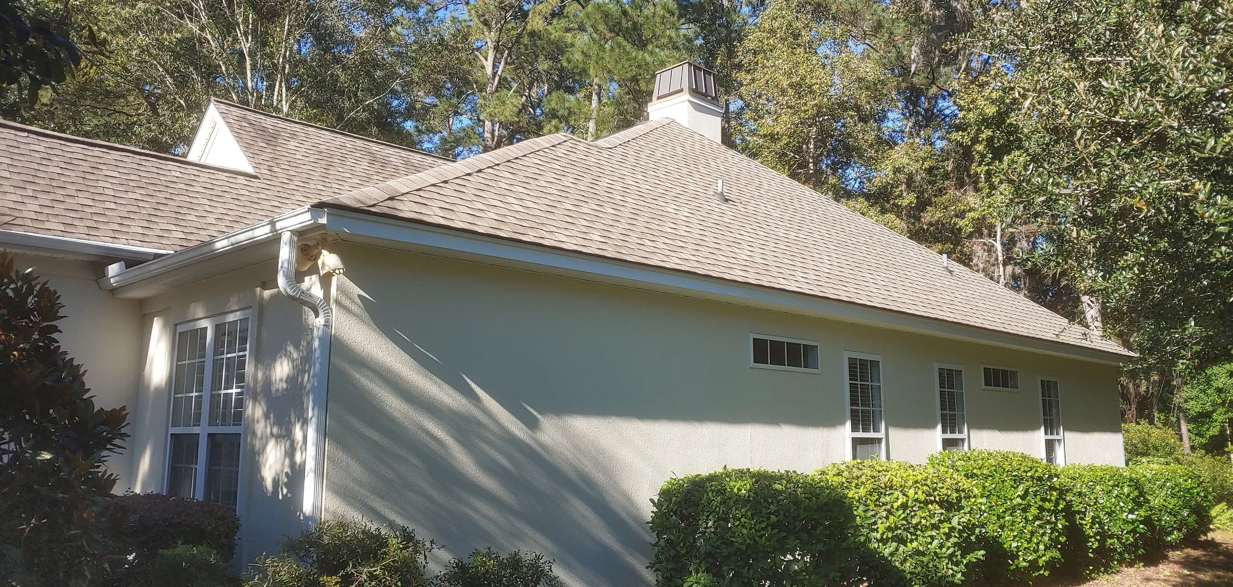 Exterior of a white house with a brown shingled roof, multiple windows, surrounded by green bushes and trees, with sunlight casting shadows.