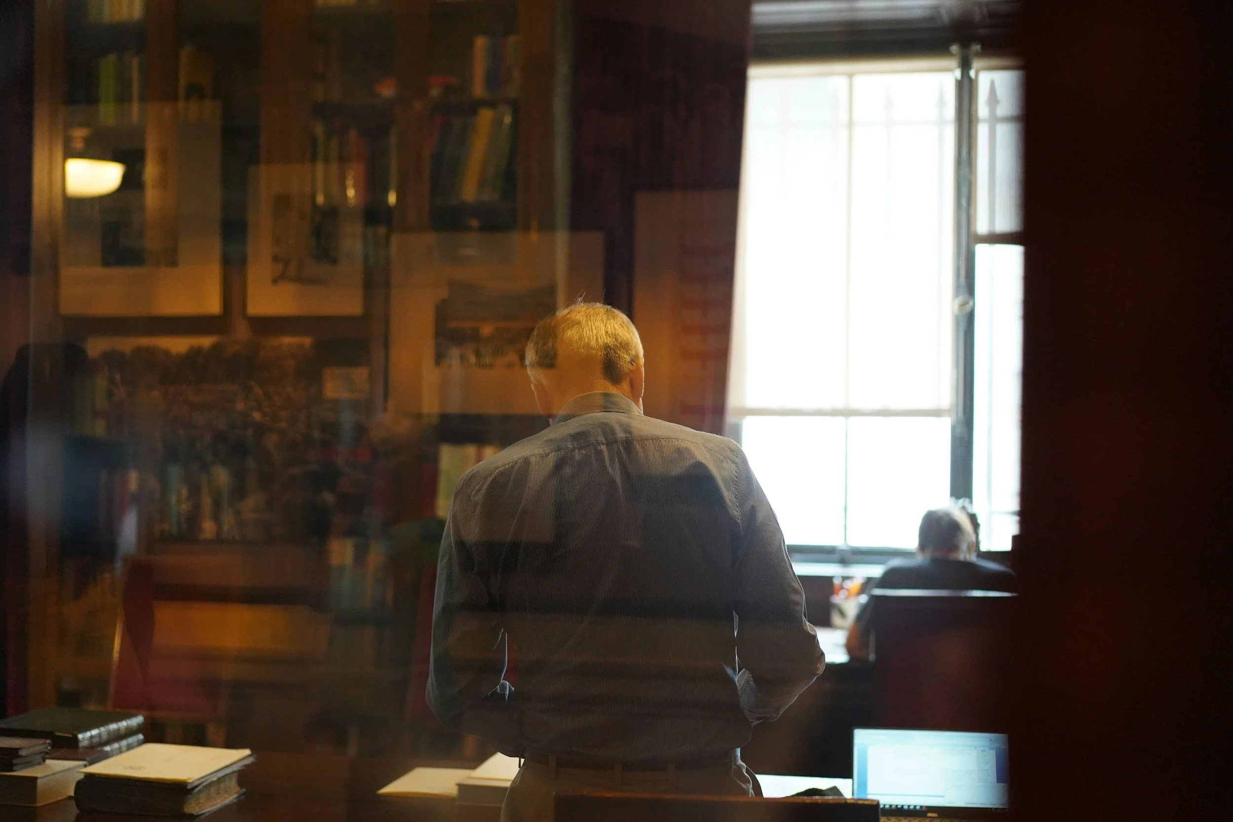 A person with light brown hair and gray shirt standing in front of a window in a room filled with books and papers, viewed through a glass window.