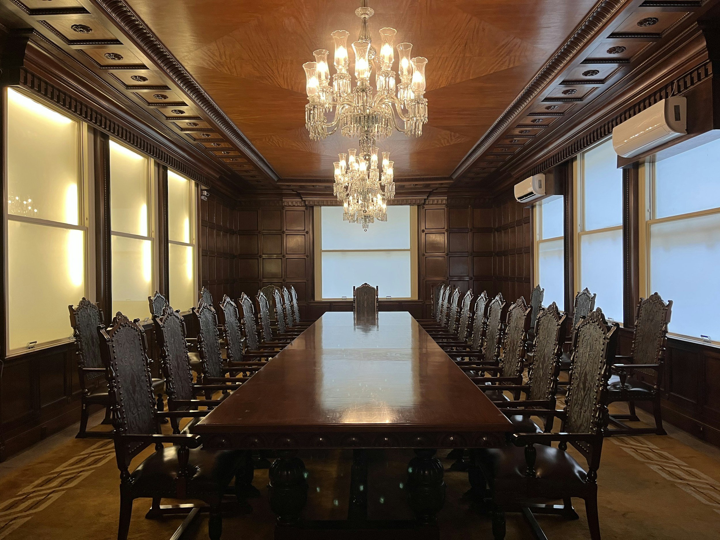 A large conference room with a long wooden table surrounded by ornate wooden chairs. The room has wood-paneled walls, a wooden ceiling, and two chandeliers hanging from the ceiling. There are large windows with shades and air conditioning units on the walls.