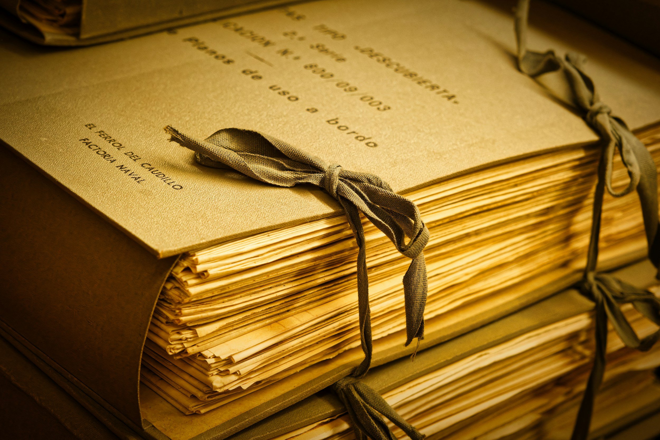 Close-up of a thick stack of aged, yellowed paper documents tied with fabric straps, with a partially visible vintage book on top.