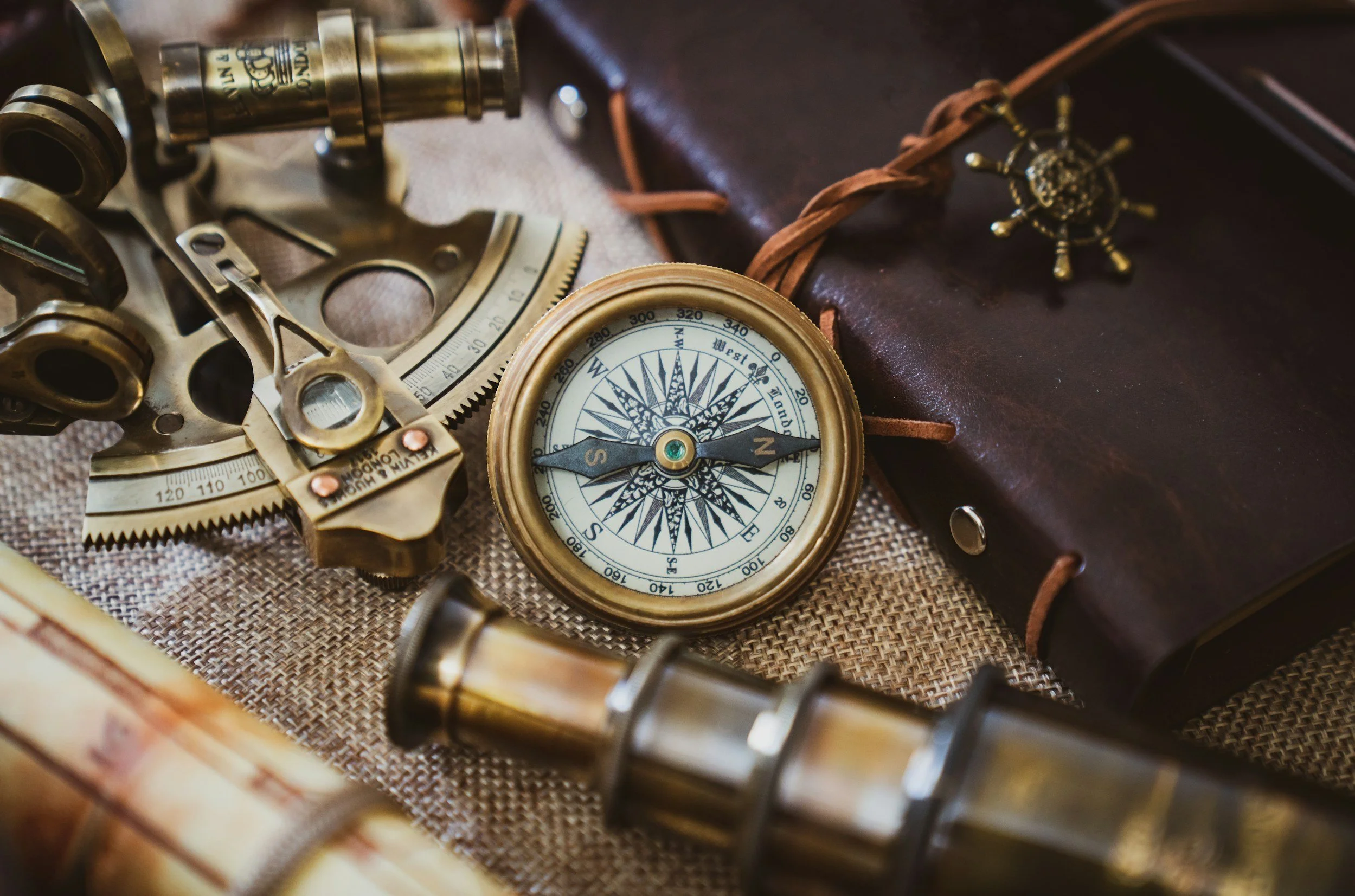 A vintage brass compass, a brass sextant, a leather notebook with a nautical-themed tie clip, and some rolled-up currency on a textured fabric surface.