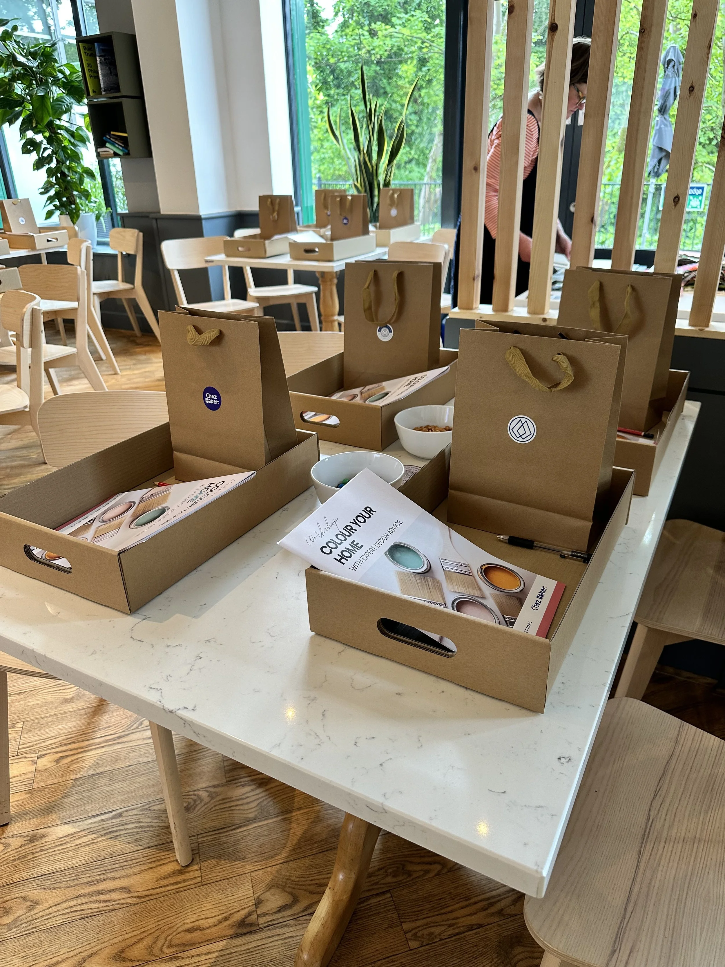 Table with cardboard gift bags and brochures in a cozy cafe with wooden floors, chairs, and large windows showing greenery outside.