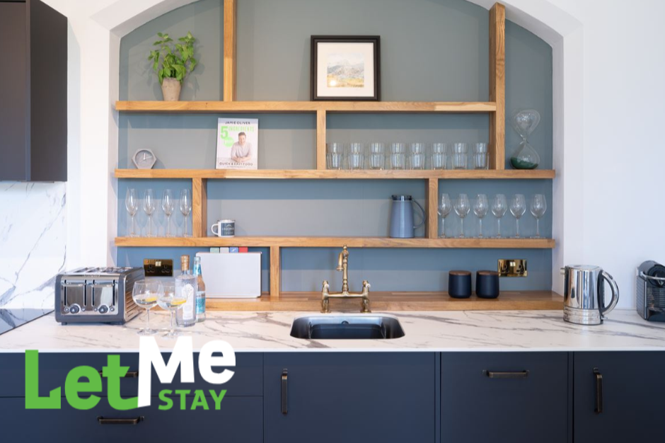 Let Me Stay help holiday home owners maximise their property's potential, offering a management service. Kitchen countertop with a sink, glassware on wooden display shelves. Home Interiors & Renovation Exhibition 2026.   #hirecumbria