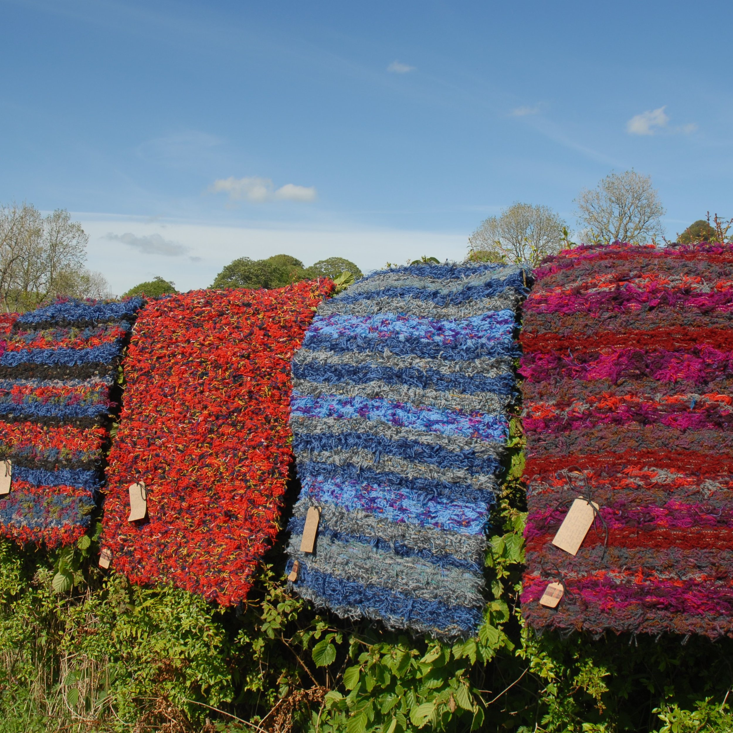 Rows of colourful woven Gneiss Rugs, handmade at Fairfield Mill, Sedbergh, hanging outdoors on a sunny day with a blue sky and trees in the background.