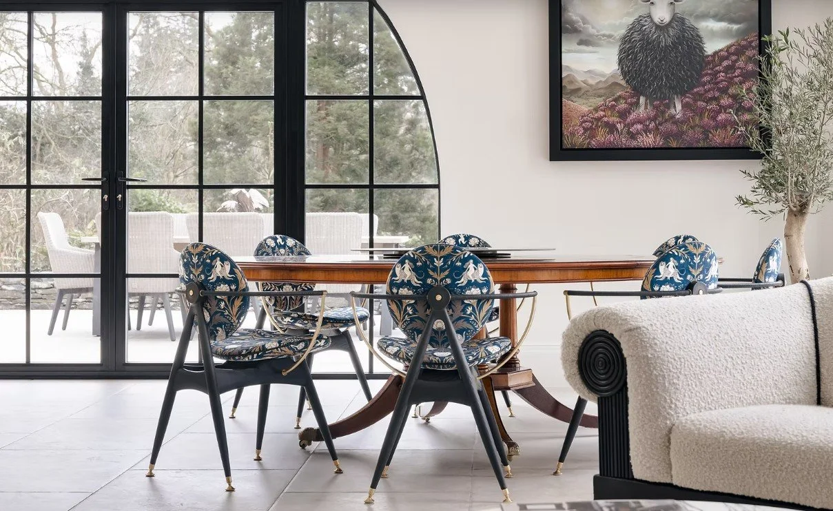 A modern dining room with a wooden oval table surrounded by six blue patterned chairs featuring unicorns and plants. Large glass doors lead to an outdoor patio with white chairs. A framed sheep artwork hangs on the wall, and a potted plant stands in 