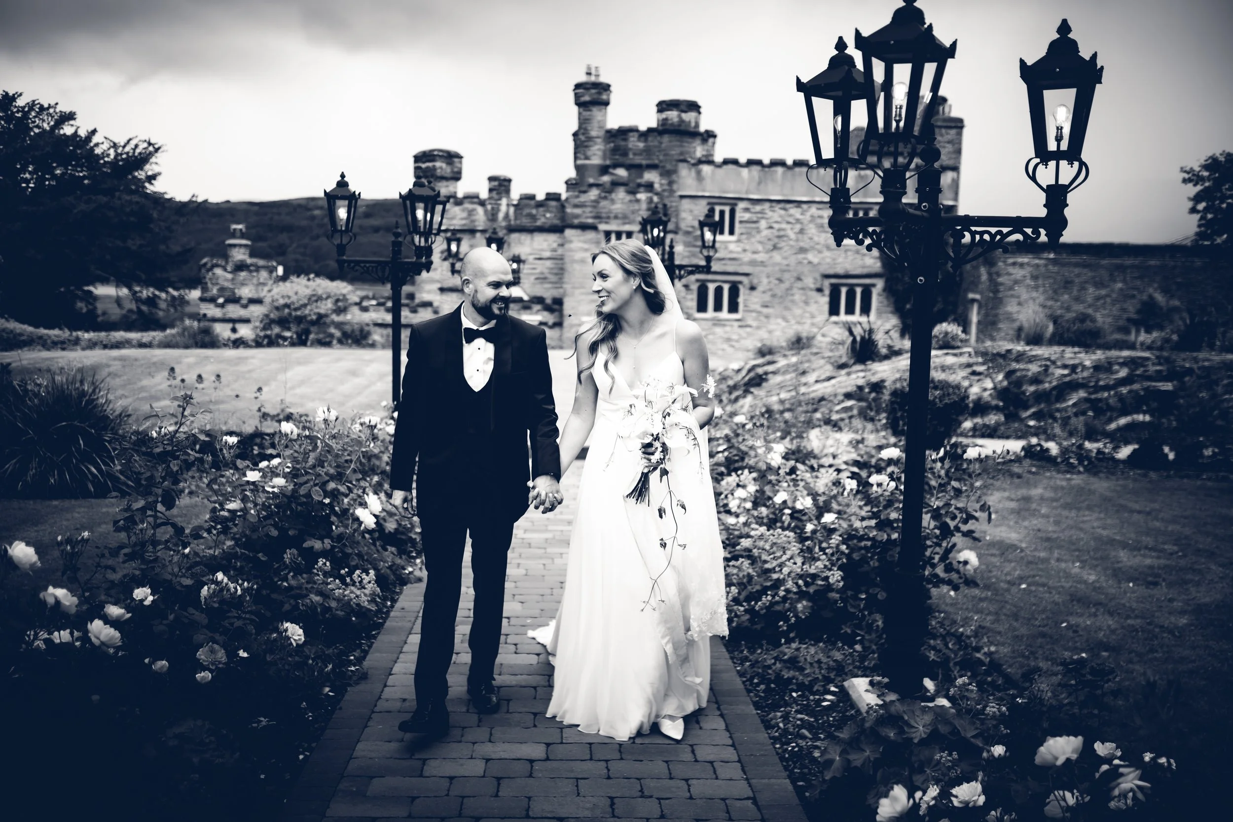 A black and white photo of a newlywed couple walking hand in hand on a brick pathway outside a castle-like building, surrounded by flowers and street lamps.
