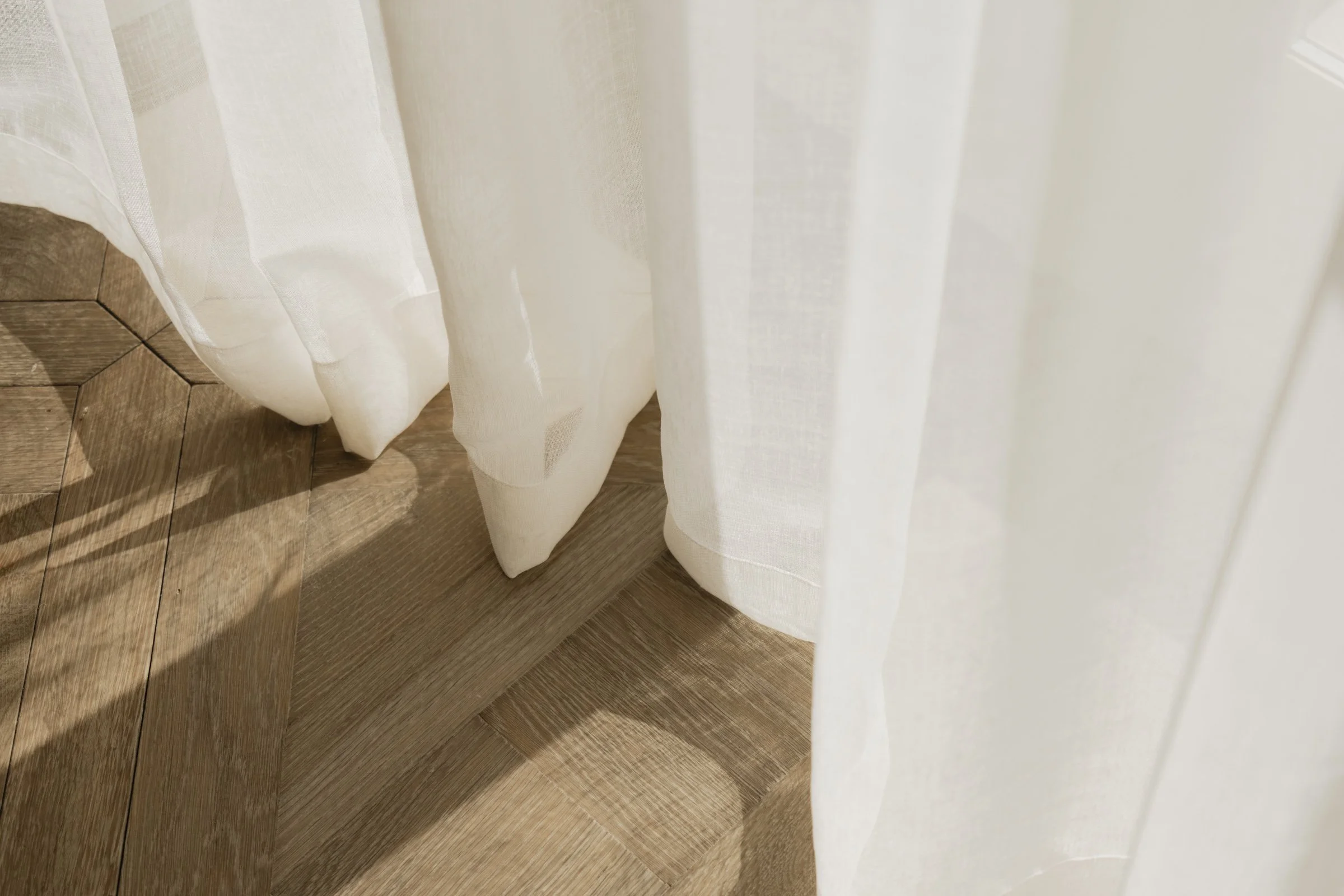 Sunlight filtering through sheer white curtains, casting shadow on wooden floor.