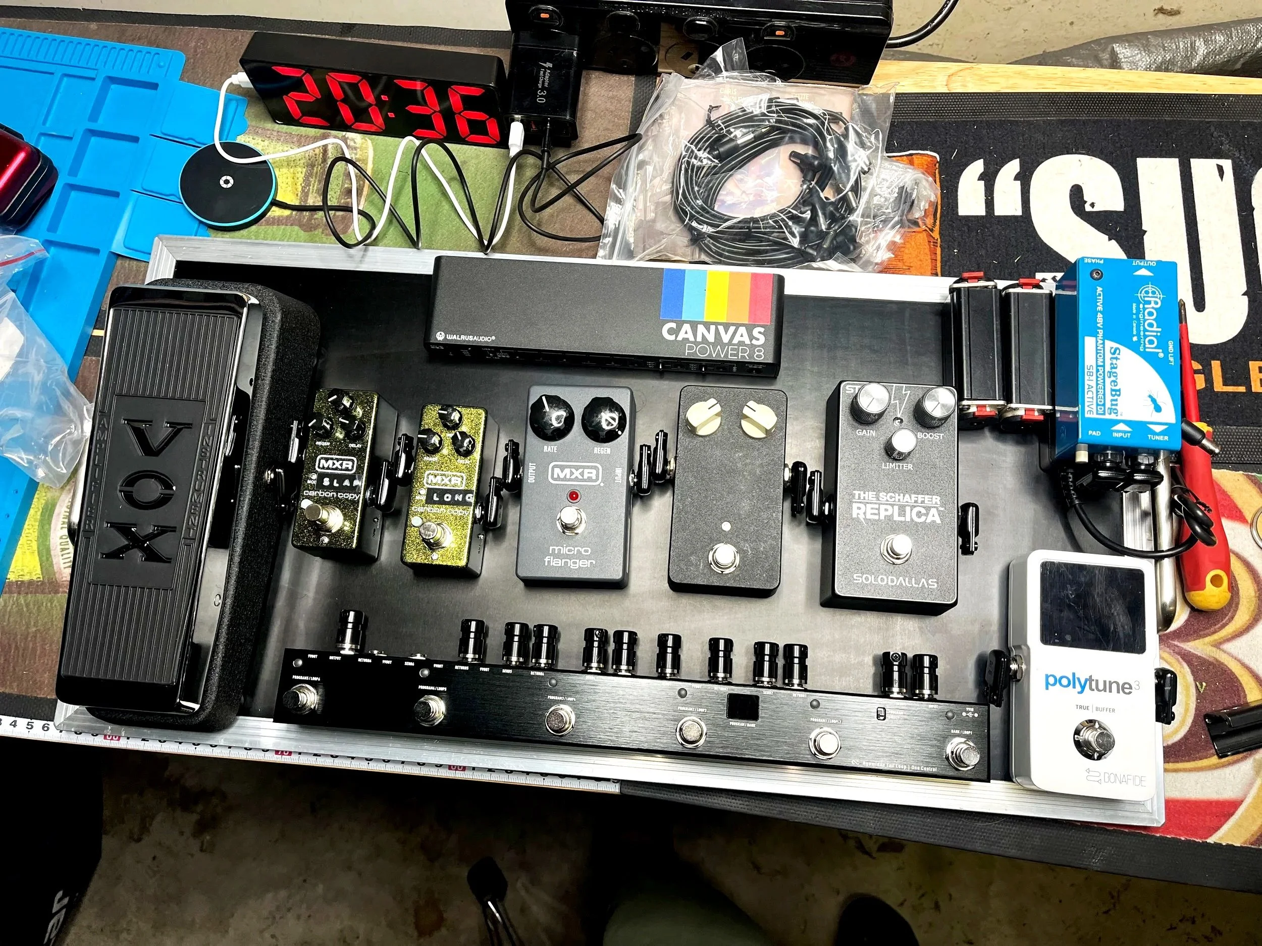A desk setup with various music production equipment including guitar pedals, a pedalboard, a digital clock displaying 20:36, a power strip, cables, and audio devices on a work surface.