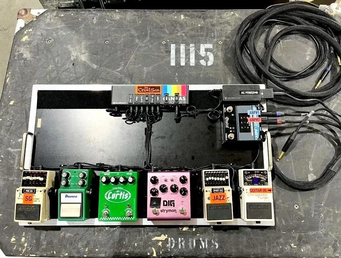 Guitar pedalboard with various effects pedals, cables, and a power supply, set up on a black surface with white markings.