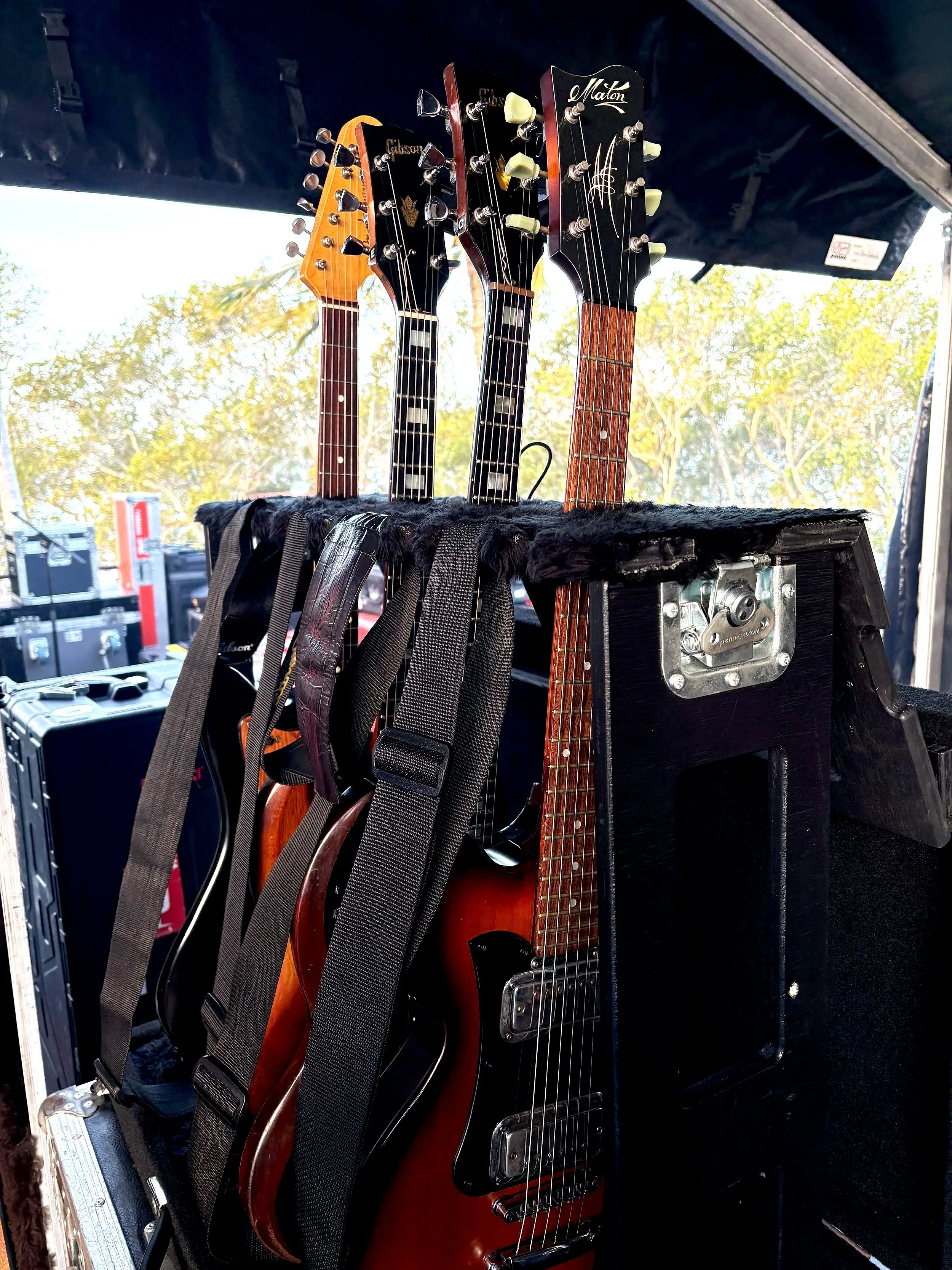 Four electric guitars on a stand with straps, placed in a black case, with a background of trees and sky.