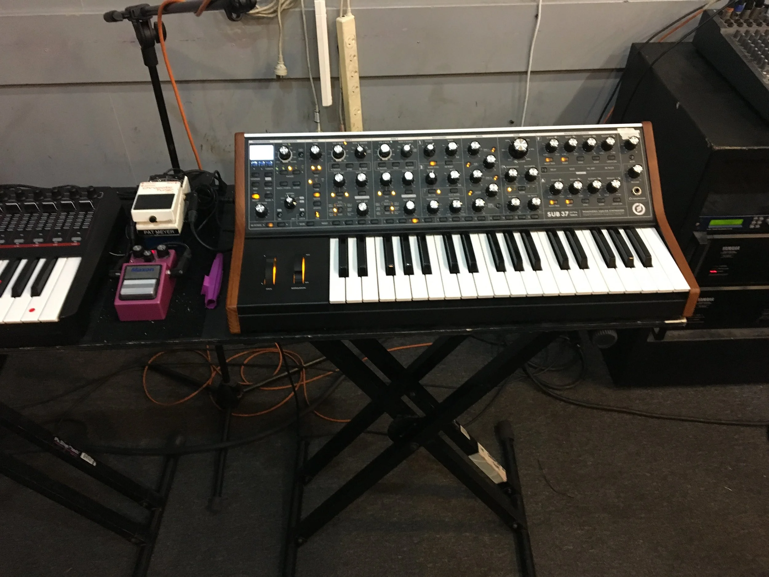 Table with musical equipment including a synthesizer with many knobs and keys, a sampler, and a mixing console in a dimly lit room.