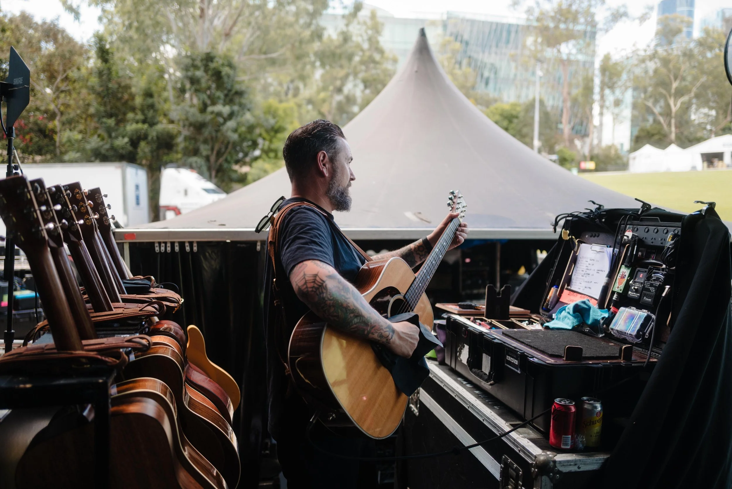 A man with tattoos on his right arm, playing an acoustic guitar at outdoor event booth with guitars on display, music equipment, and drinks in front of trees and a modern building in the background.