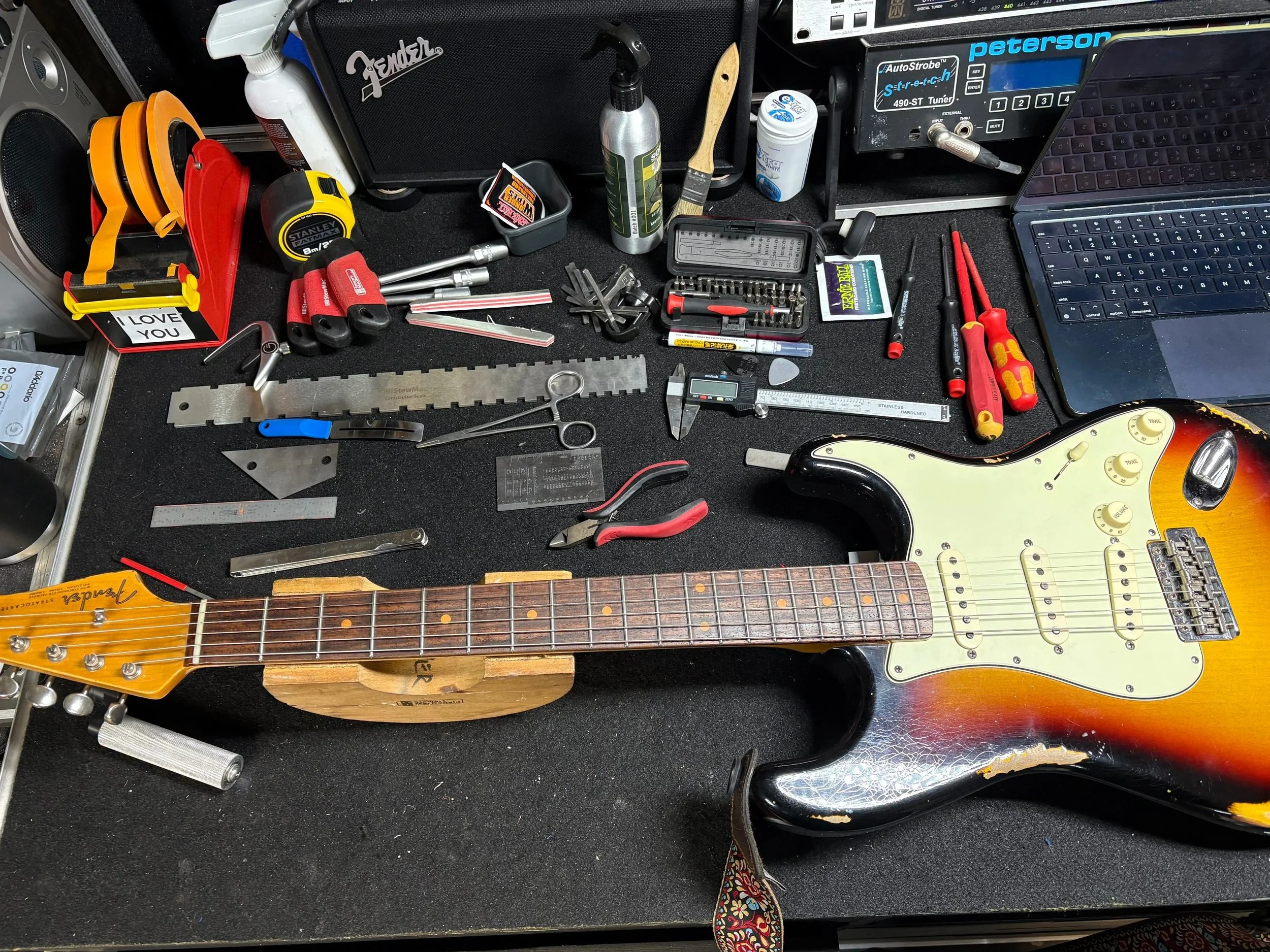 A workbench with various tools including screwdrivers, a wrench, a saw, a laser level, a digital caliper, and a guitar. There are also cleaning supplies, a laptop, and a Fender amplifier. The background shows speakers and other electronic equipment.