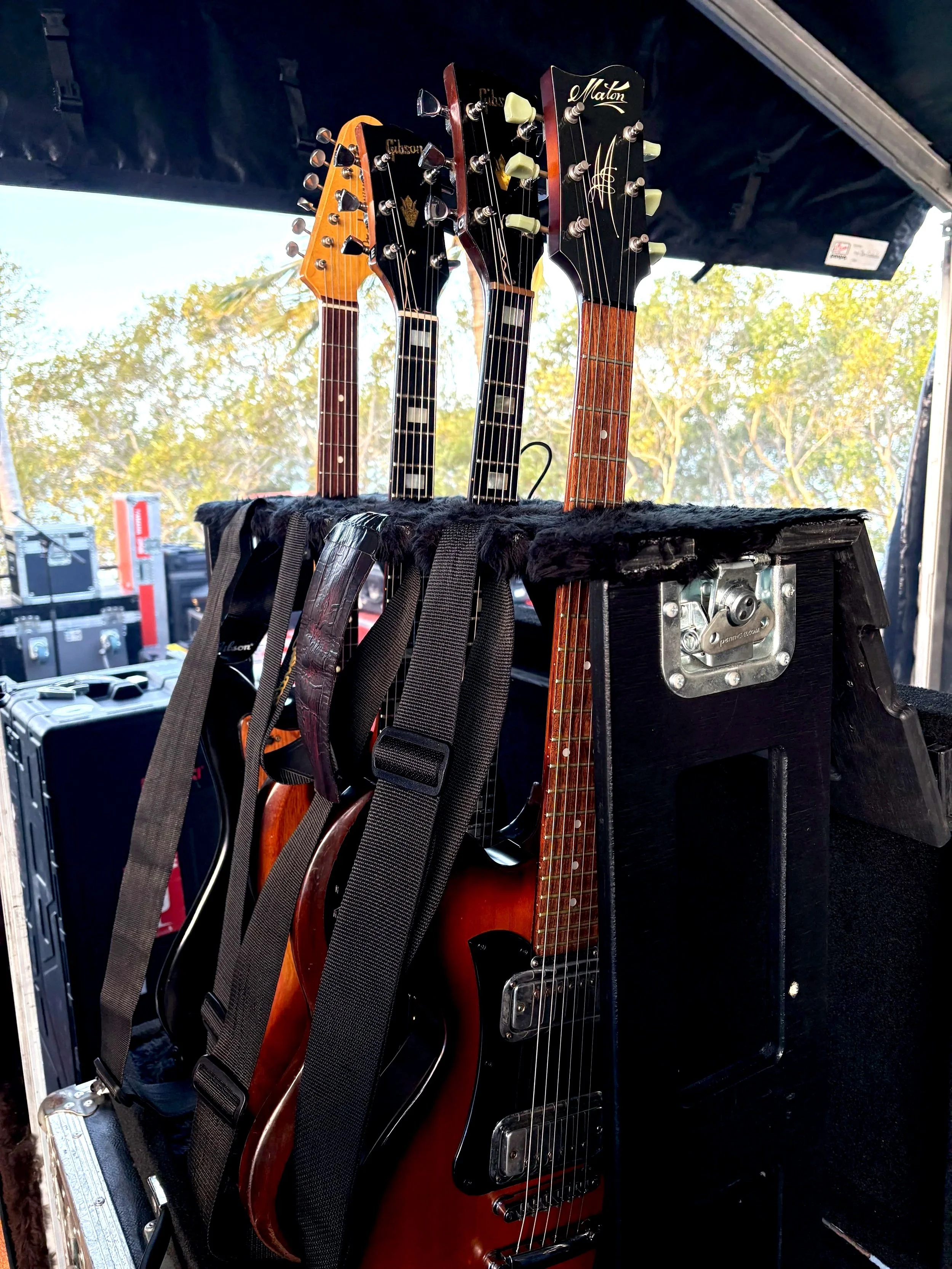 Four electric guitars hanging on a black case in an outdoor setting, with trees and sky in the background.