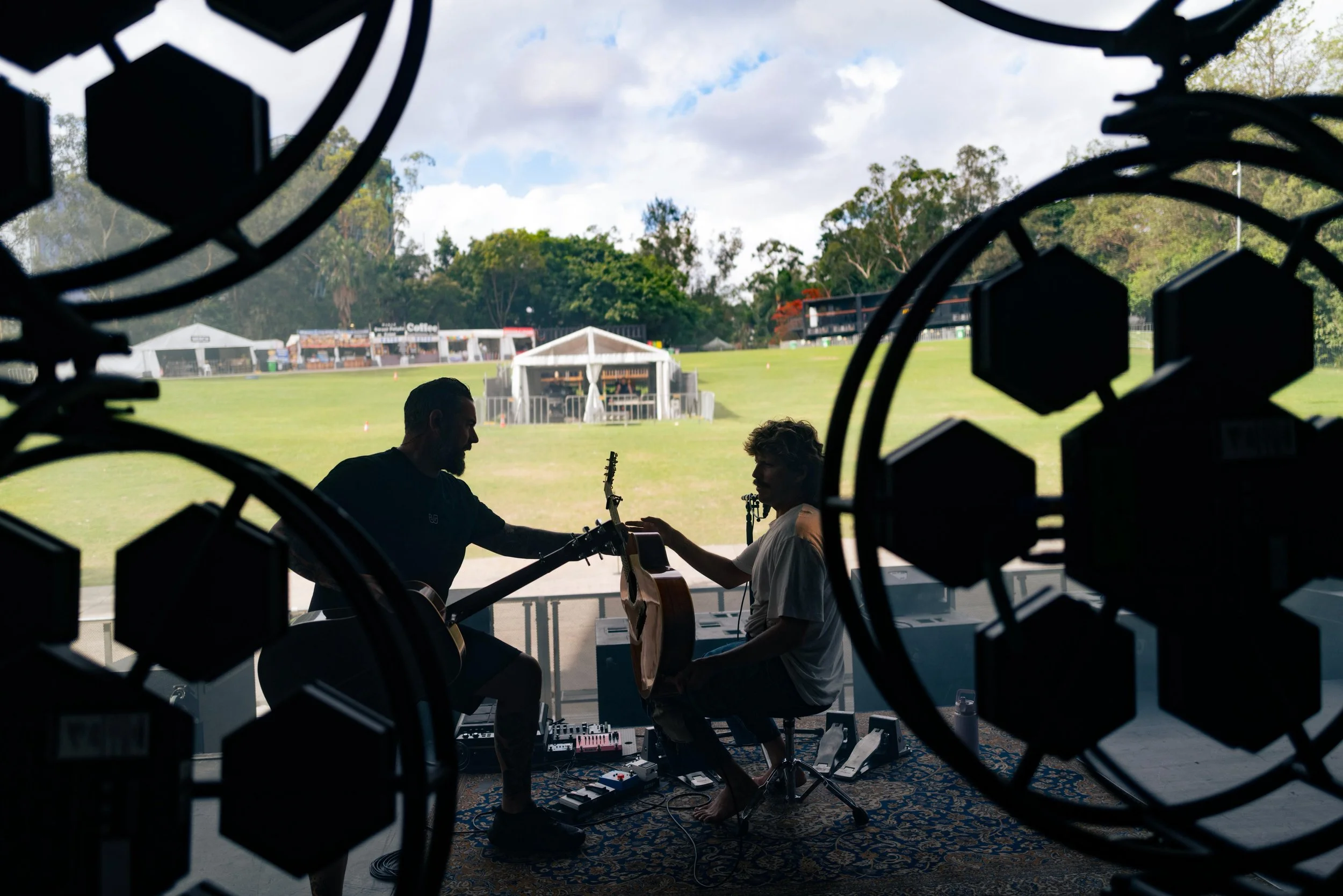 Two musicians are playing guitars on a stage outside during the daytime, viewed through a decorative fence with geometric patterns.