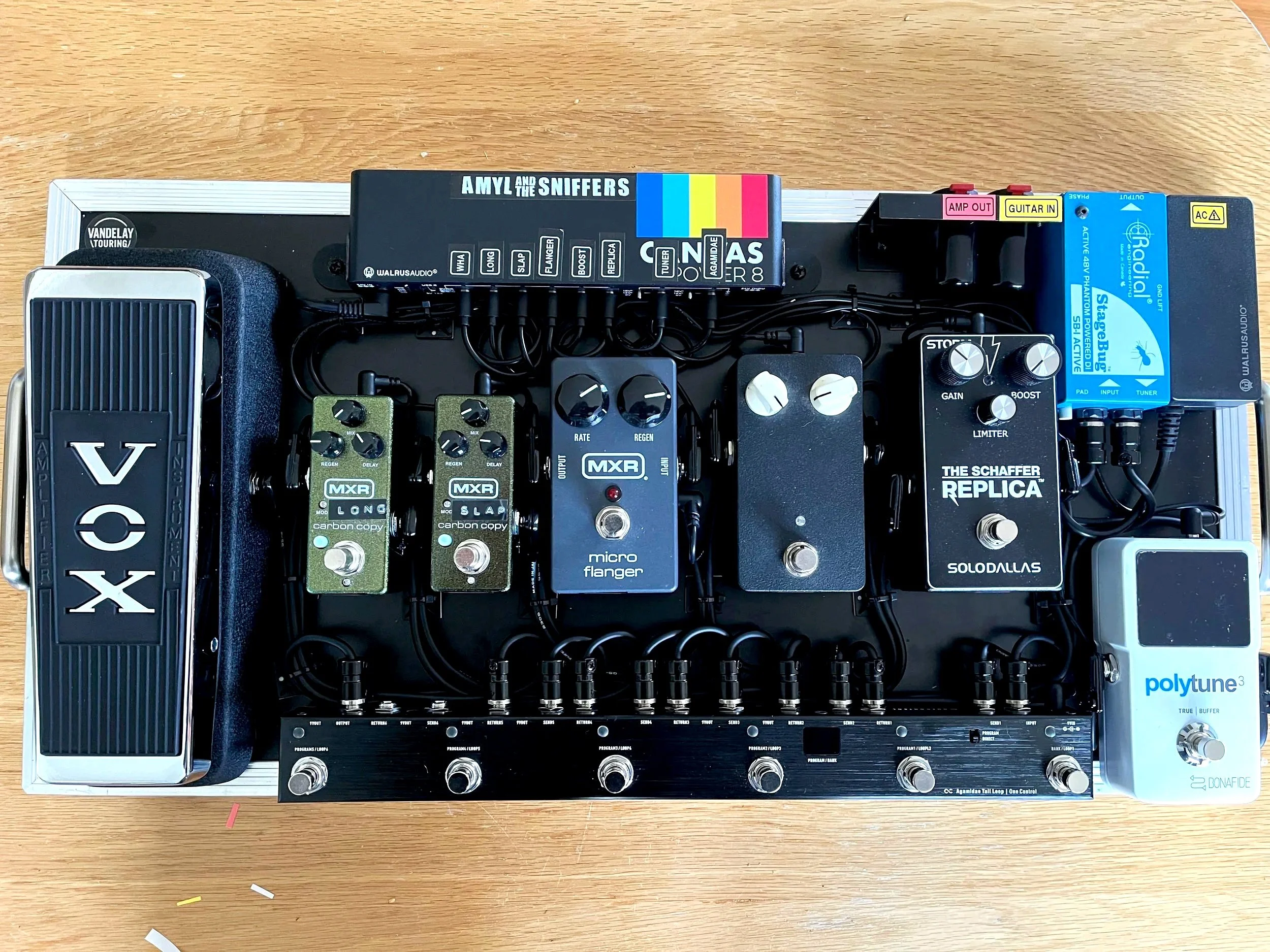 An overhead view of a music pedalboard with various guitar effects pedals and a digital tuner, on a wooden surface.
