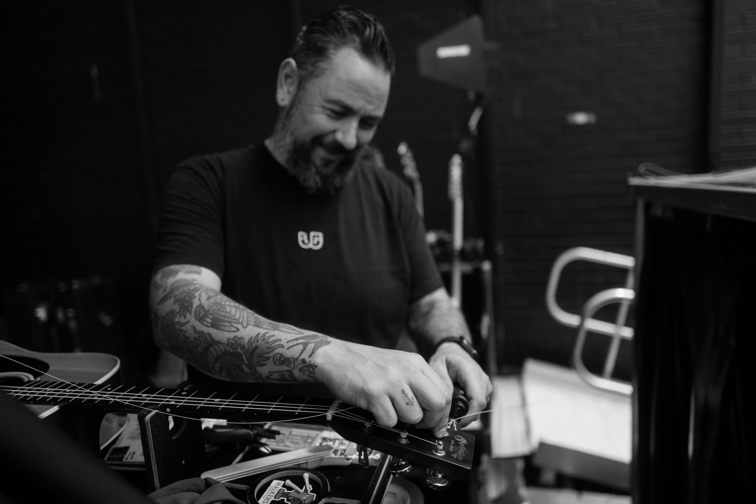 Black and white photo of a man with tattoos on his arm, playing a guitar and smiling.