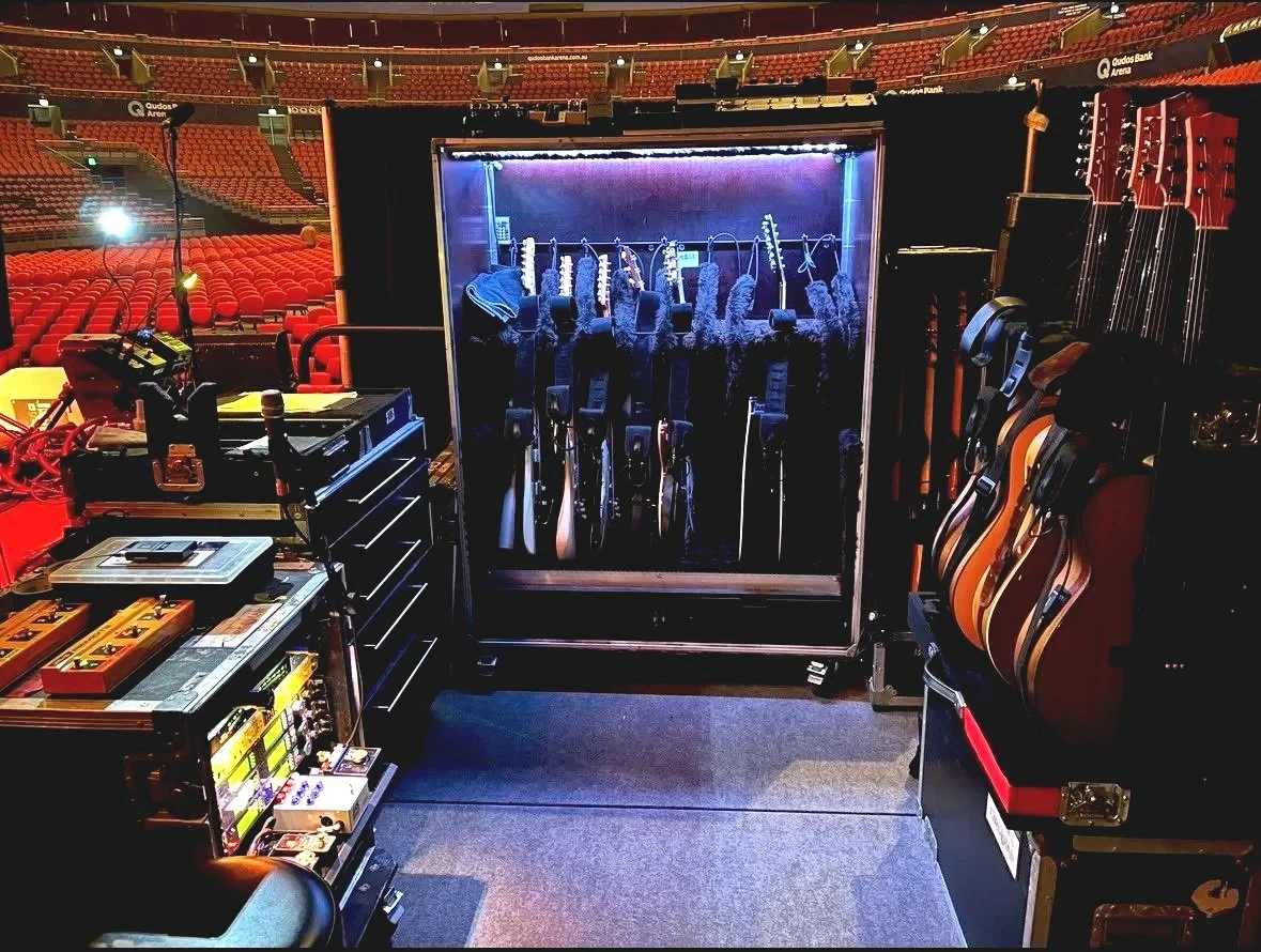 A stage setup with guitars and audio equipment, including a rack of guitars behind a glass door, musical instruments on the right, and audio gear on the left, in a large arena with red seating.