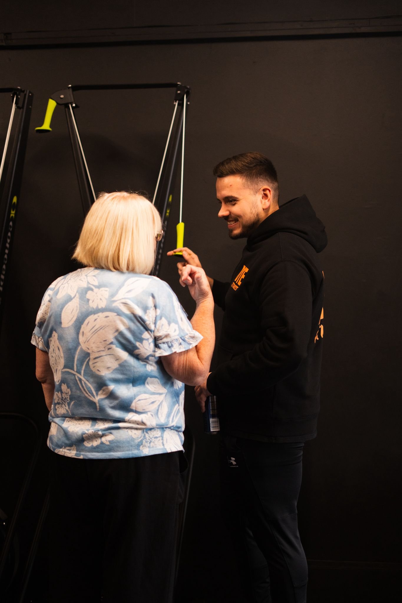 An elderly woman with blonde hair and a blue floral blouse talking to a young man in a black hoodie, standing in front of a black wall with workout equipment.