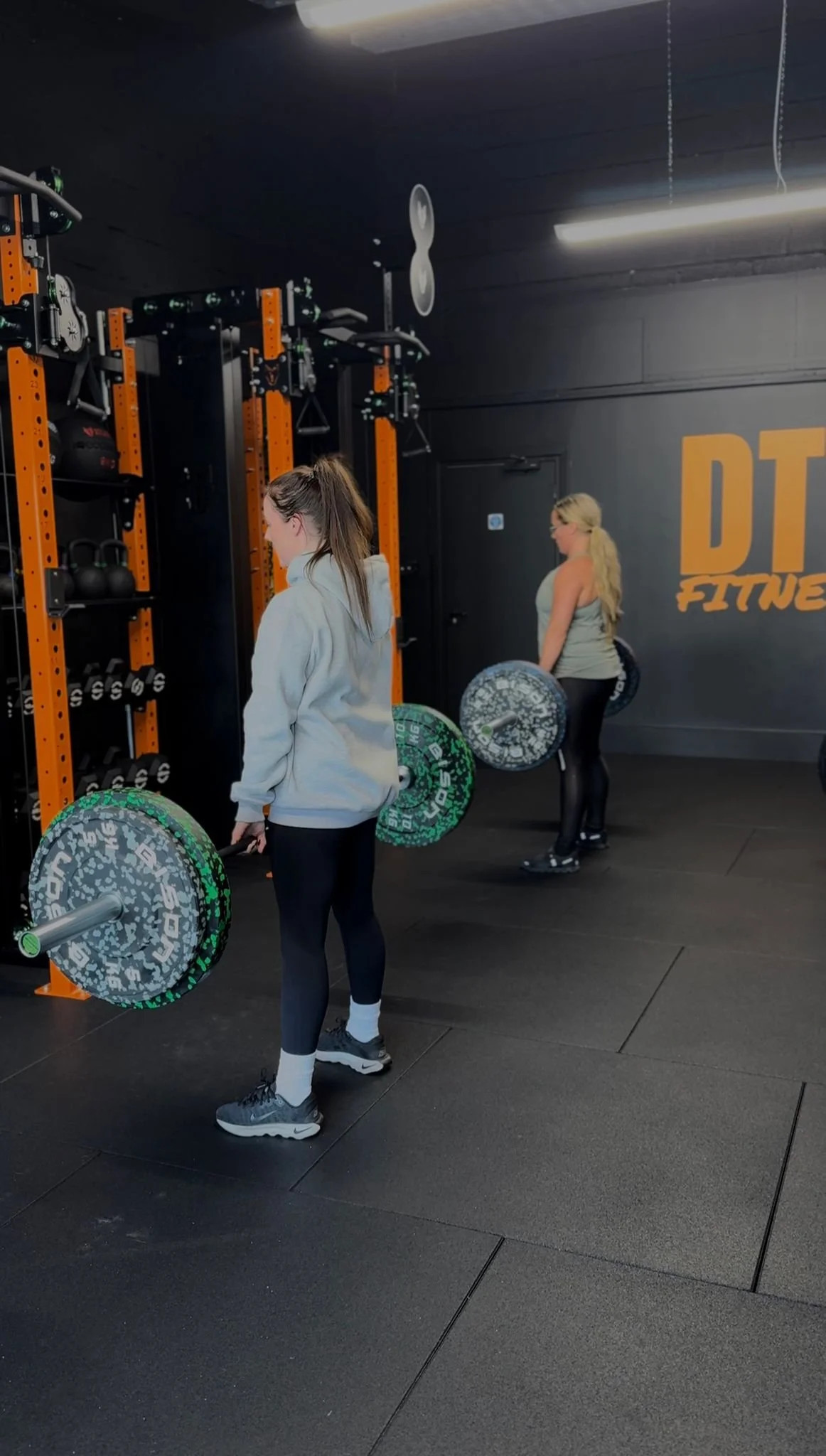 Two women lifting barbells in a gym with black walls and rubber flooring. One is in the foreground wearing a gray hoodie and black leggings, and the other is in the background wearing a gray tank top and black leggings.