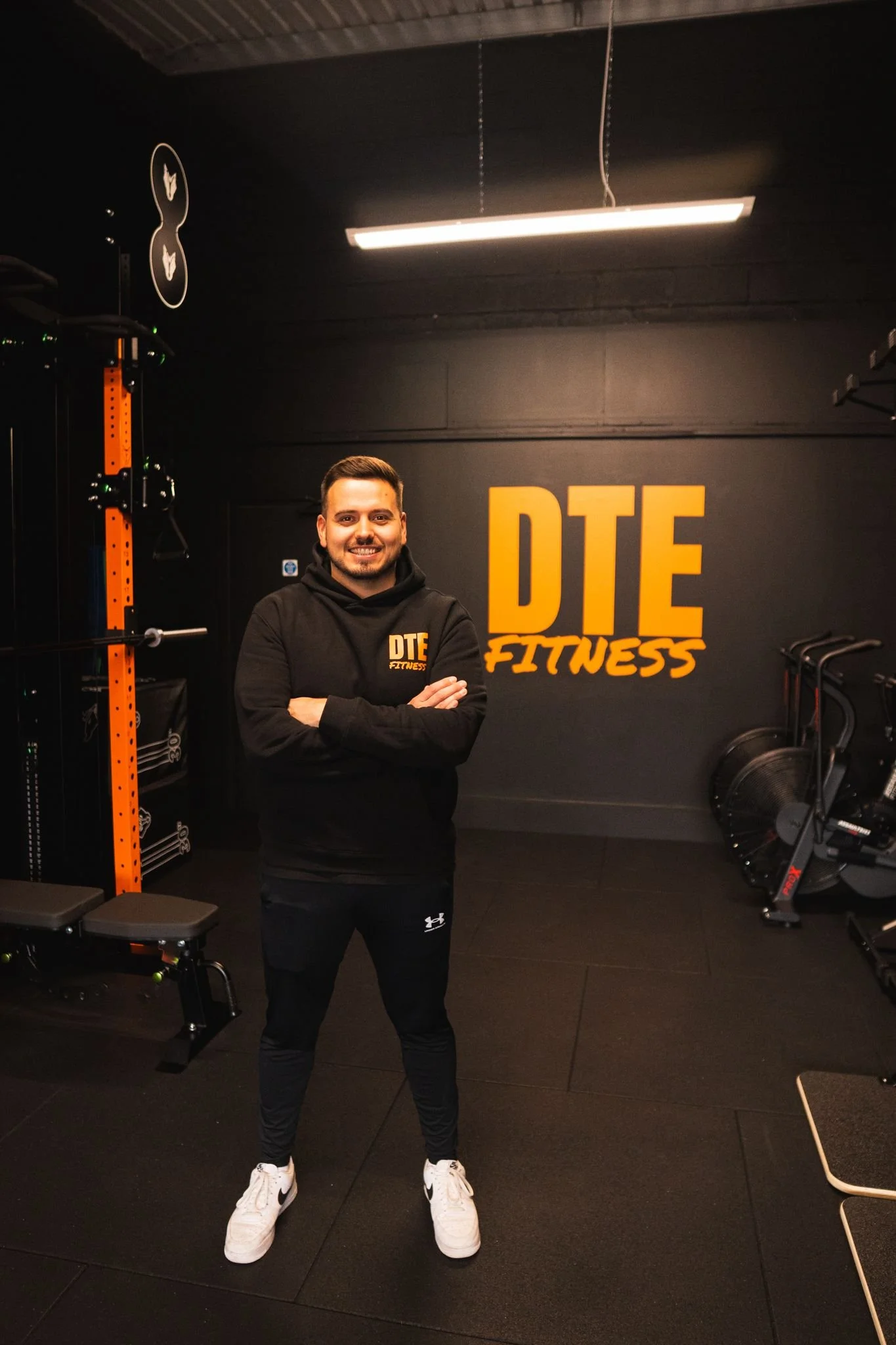 A man stands with arms crossed in a gym with black walls, wearing a black hoodie and black pants, in front of a yellow and black sign that reads "DTE Fitness."