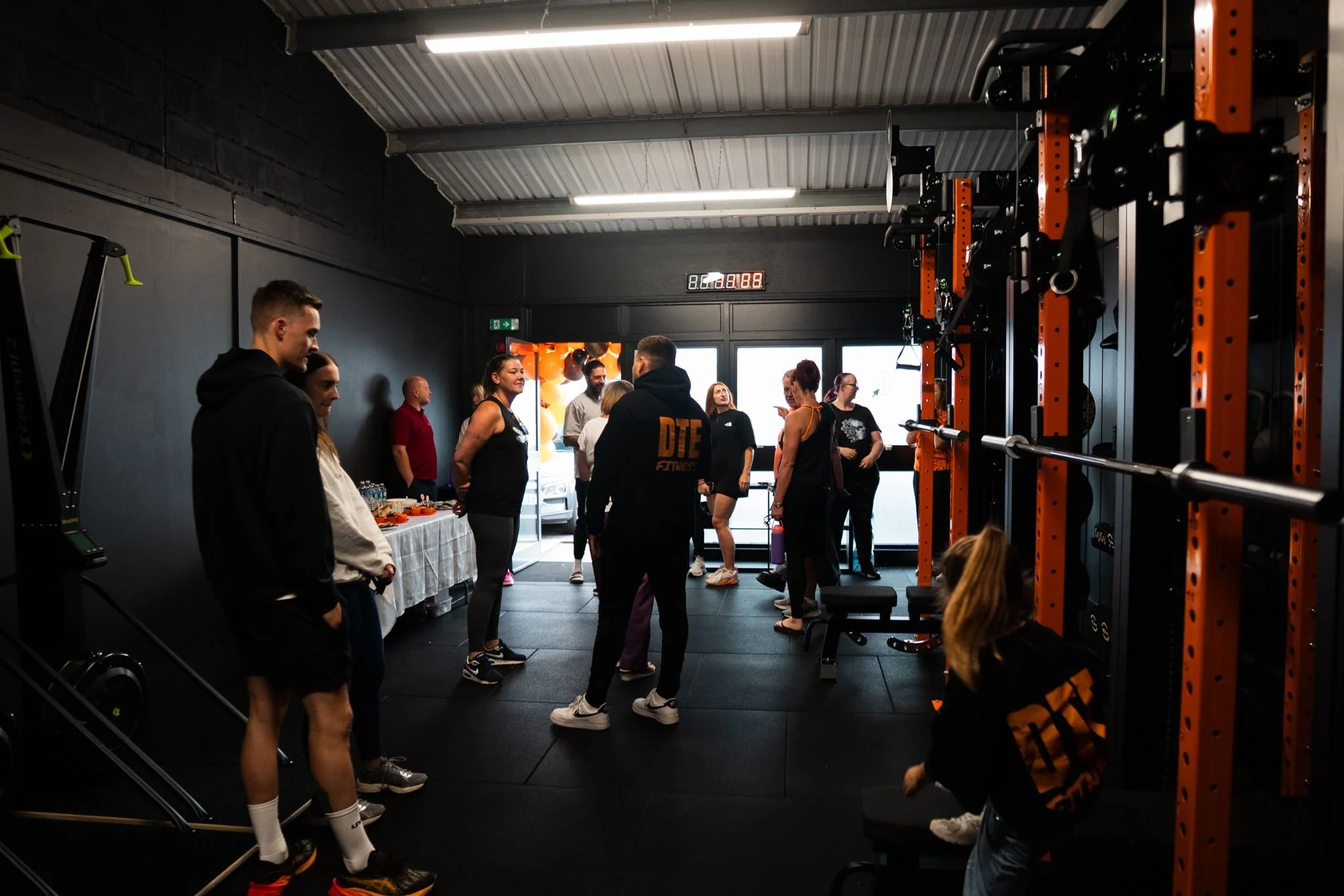 People gathered in a gym near orange and black weightlifting equipment, some talking and others sitting or standing, with a table of snacks or drinks on the side.