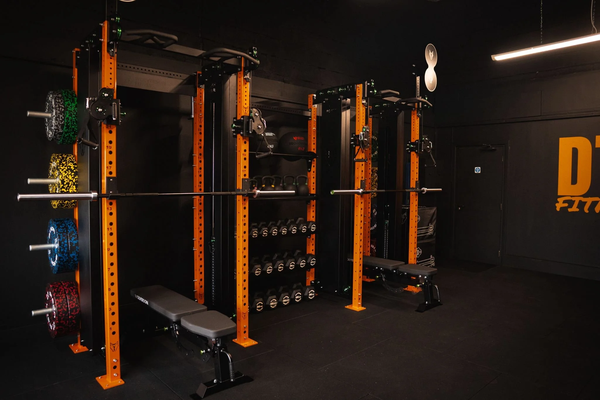 Empty gym with orange and black workout equipment including weight racks, barbells, dumbbells, and a weight bench.