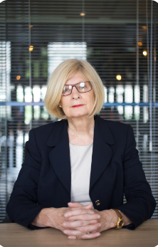 An older woman with blonde hair, glasses, and a serious expression, sitting at a table in a modern office or conference room with glass walls and reflective surfaces.