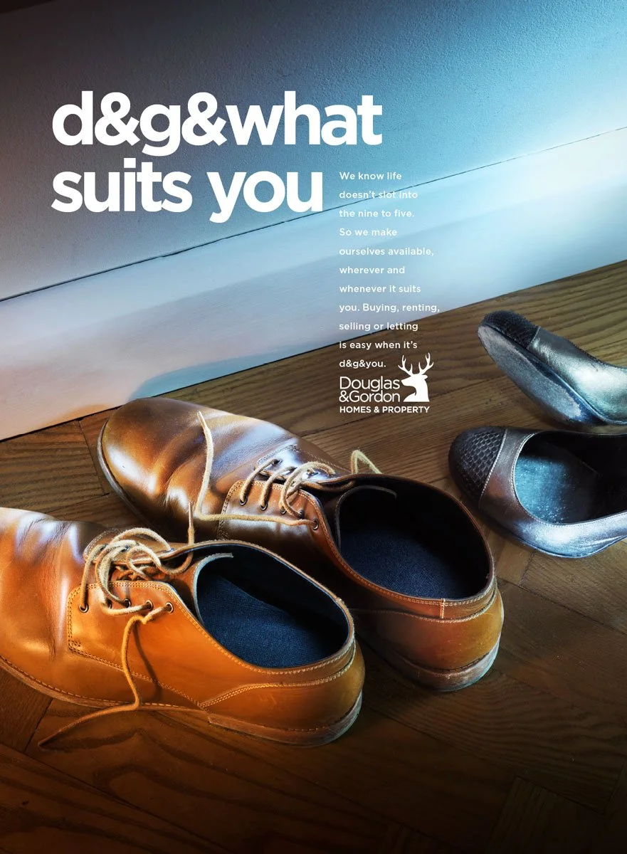 Une paire de chaussures en cuir marron et deux paires de chaussures en cuir noir, toutes posées sur un plancher en bois, à côté d'une boîte ou panneau comportant un texte promotionnel pour Douglas & Gordan HOMES & PROPERTY.
