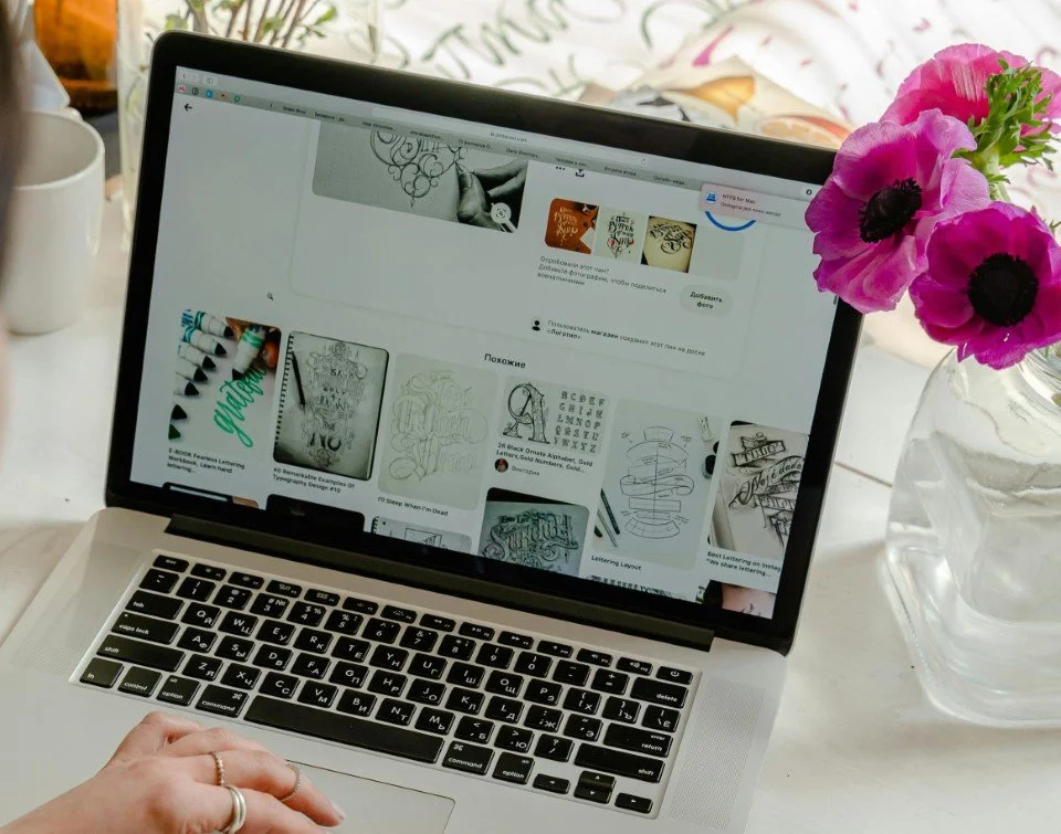 Un ordinateur portable affichant des images et des idées de design, posé sur une table blanche à côté d'un vase avec des fleurs roses.