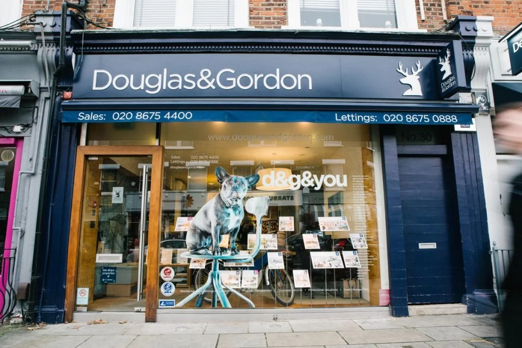 Vitrine d'une agence immobilière 'Douglas & Gordon' avec un panneau de contact, une affichage de vente et de location, une mascotte en forme de chien sur une chaise et des affiches promotionnelles dans la fenêtre.