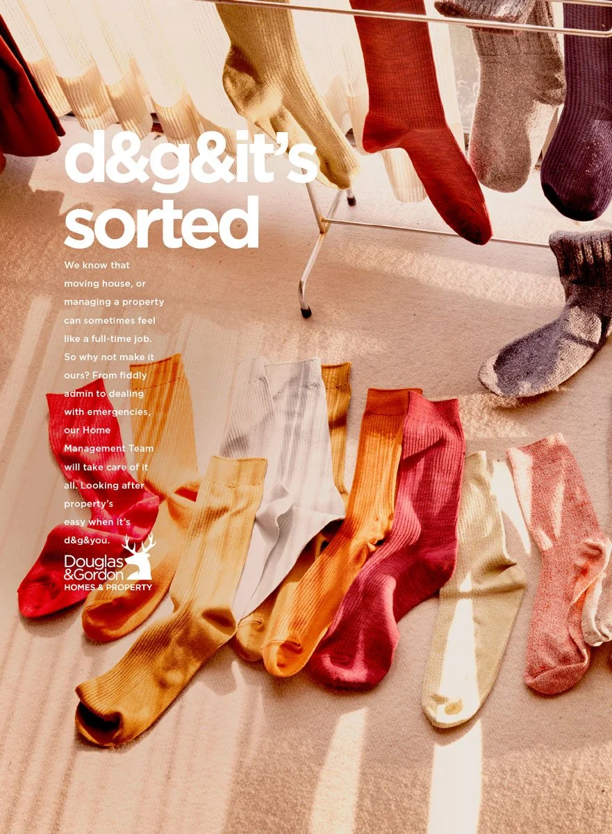 Une rangée de chaussettes colorées suspendues sur une corde, avec des coussins dans des tons ambre, bordeaux, gris, noir, gris clair, beige et rose pâle, éclairées par la lumière naturelle