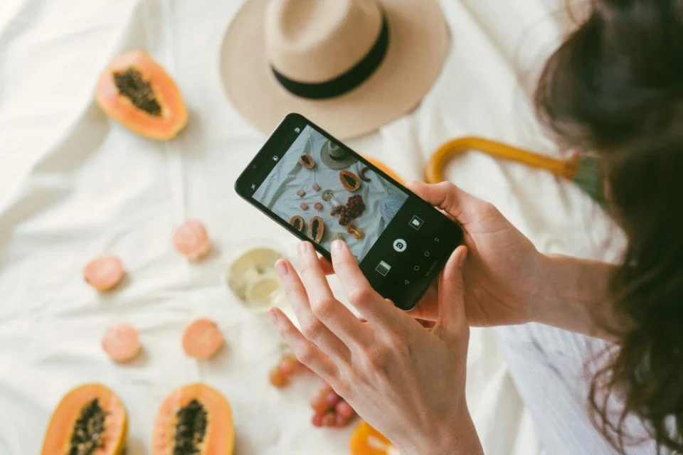 Une personne prend une photo de fruits et de macarons avec son smartphone sur une nappe blanche.